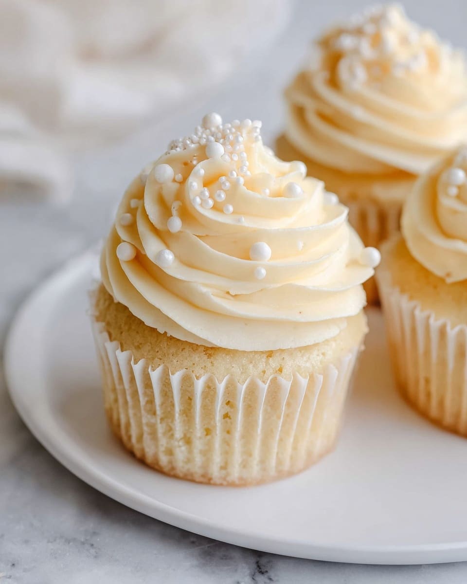 The image shows a close-up of a vanilla cupcake on a white plate, placed on a white marbled surface. The cupcake has one main layer of light golden cake at the bottom, topped with a thick swirl of creamy, pale yellow buttercream frosting. The frosting is decorated with small white sugar pearls and tiny white sugar crystals scattered on top, adding texture and contrast to the smooth frosting. Two more cupcakes are slightly visible in the background, also topped with similar creamy frosting and decorations. Photo taken with an iphone --ar 4:5 --v 7