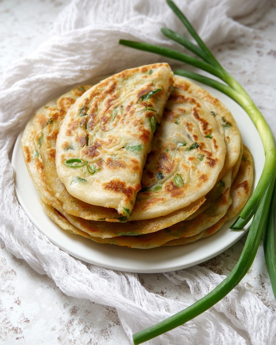 The image shows a stack of four round flatbreads on a white plate, each flatbread thin with golden-brown spots and light green herbs mixed inside the dough. The top flatbread is slightly folded at the edge, revealing its soft texture inside. The flatbreads have a slightly uneven surface with visible layers and crispy edges. On the right side of the plate, there are a few green spring onion stalks resting on the white marbled texture. The overall setting is bright and soft, with cream-colored cloth parts visible around the plate. photo taken with an iphone --ar 4:5 --v 7