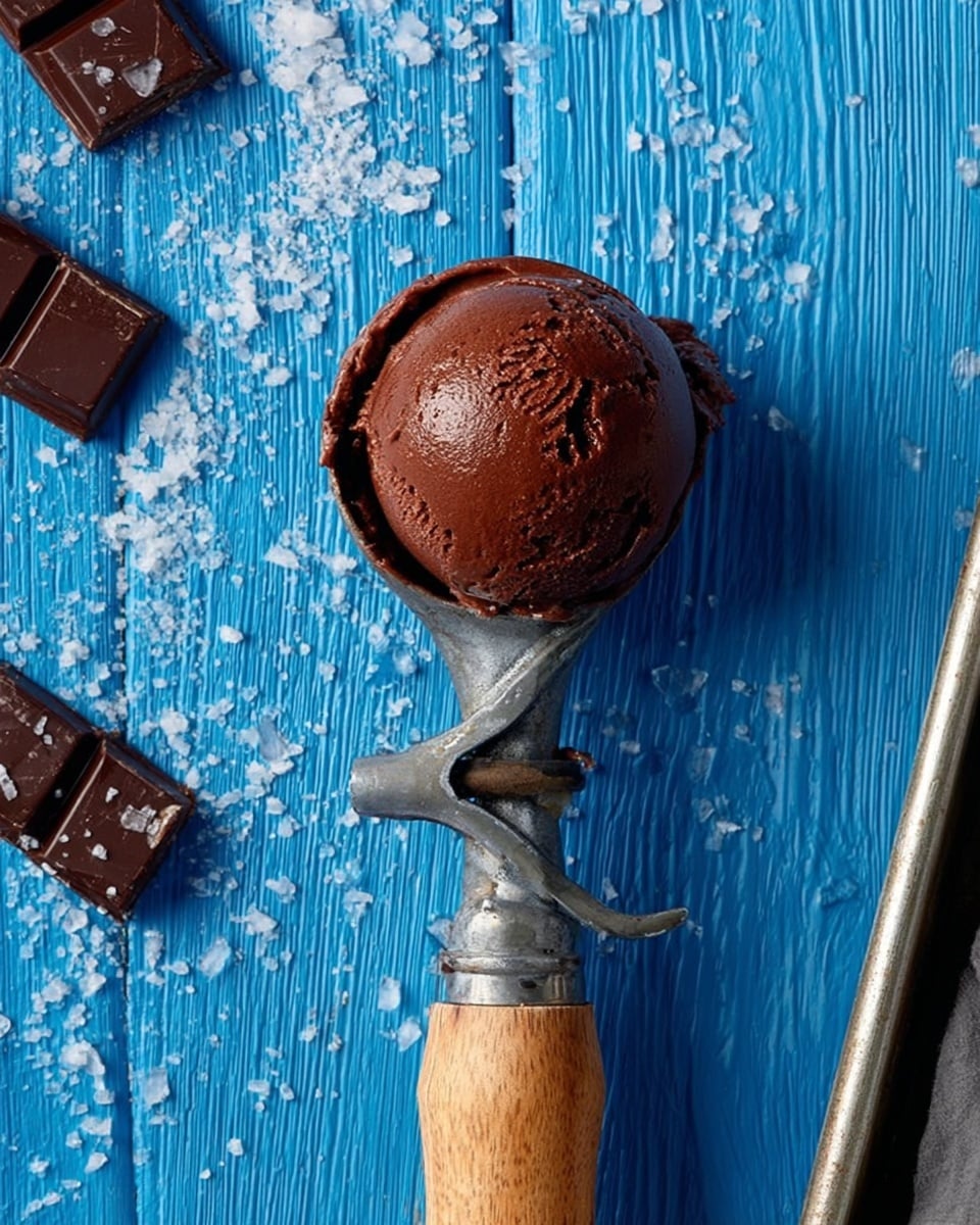 A metal ice cream scoop with a wooden handle holds a single round scoop of rich dark brown chocolate ice cream, showing a slightly shiny and smooth texture on the surface. The scoop is centered over a white marbled wooden surface that has scattered grains of coarse white sea salt. Pieces of broken dark chocolate squares are placed on the upper left side of the scene, and the top right corner shows the edge of a metal baking pan visible. The overall color contrast is strong between the dark chocolate and the bright blue wooden background with subtle wood grain patterns. photo taken with an iphone --ar 4:5 --v 7