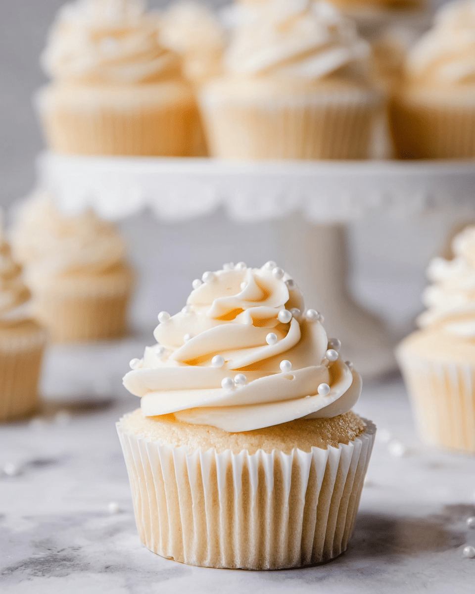A single vanilla cupcake sits in the front, with a light golden base wrapped in a ridged white paper liner. On top, there are two thick, swirled layers of creamy white frosting, decorated with small white sugar pearls scattered evenly. In the background, there is a white cake stand holding more similarly decorated cupcakes, slightly out of focus. The scene is set on a white marbled surface, giving a clean and elegant look. Photo taken with an iphone --ar 4:5 --v 7