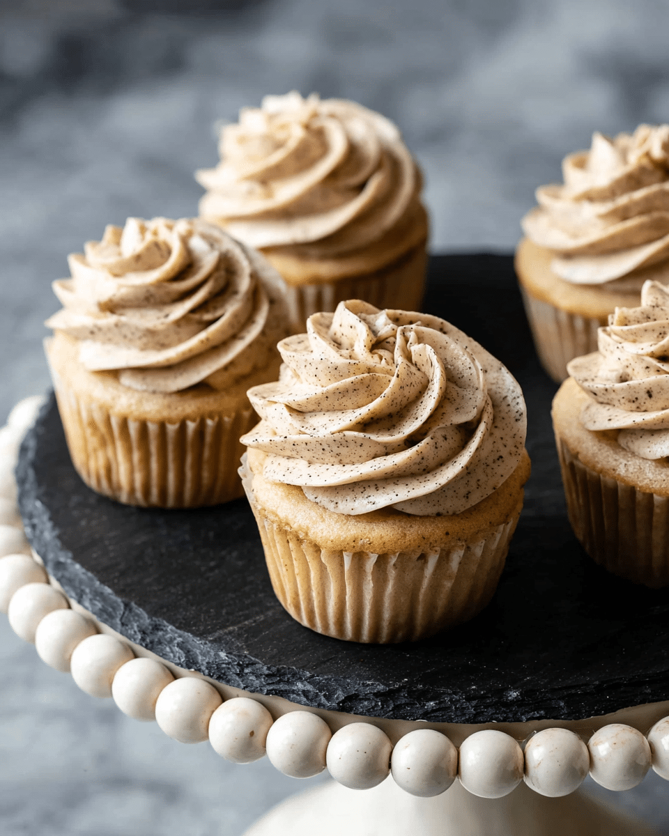 Five vanilla cupcakes each have one swirl of light brown frosting with dark specks on top, showing a smooth and creamy texture. The cupcakes sit closely on a round black slate plate, which is placed on a white marbled textured surface decorated with large, worn white beads around the edge. The cupcakes are without any additional decoration, and the frosting is neatly piped with soft curves. Photo taken with an iphone --ar 4:5 --v 7