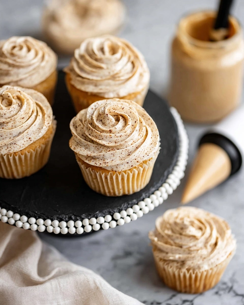 The image shows creamy, light brown frosting swirled in a rose shape on top of golden yellow cupcakes, each with a slightly textured frosting that has darker brown specks. The cupcakes are placed on a round black slate board with a decorative white beaded edge, set on a white marbled surface. In the background, there is a piping bag with similar frosting, and a jar filled with the same light brown frosting, both on the white marbled surface. A soft, off-white cloth is partially visible near the bottom left corner. photo taken with an iphone --ar 4:5 --v 7