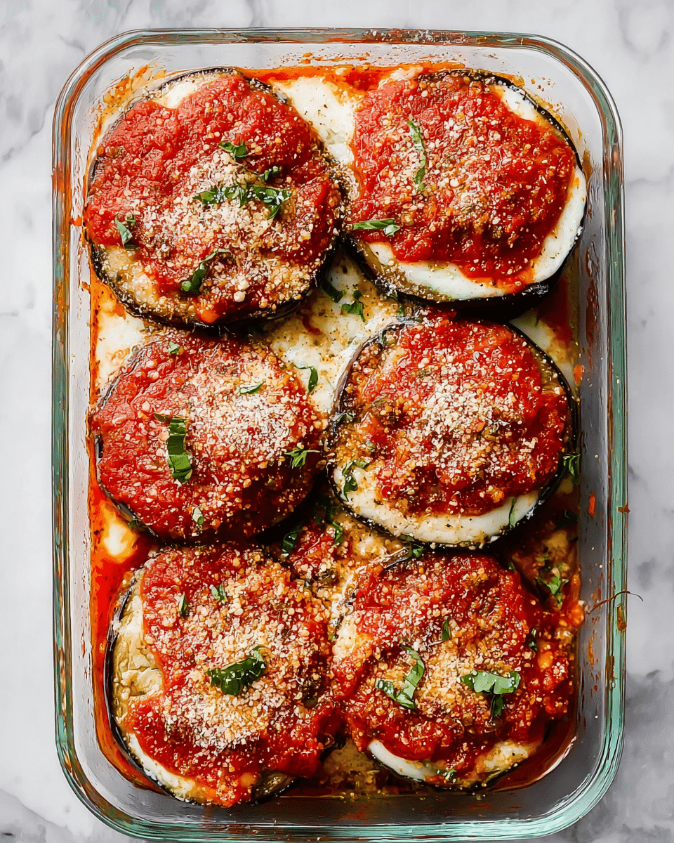 A clear glass baking dish filled with six large, round slices of eggplant layered in two visible layers. The bottom layer is creamy white melted cheese, topped with bright red tomato sauce spread thickly on the top layer of eggplant slices. The sauce is sprinkled with grated Parmesan cheese and small pieces of fresh green basil leaves scattered across. The edges show a bit of thick orange tomato sauce spread unevenly around the sides of the dish. The background has a white marbled texture. photo taken with an iphone --ar 4:5 --v 7