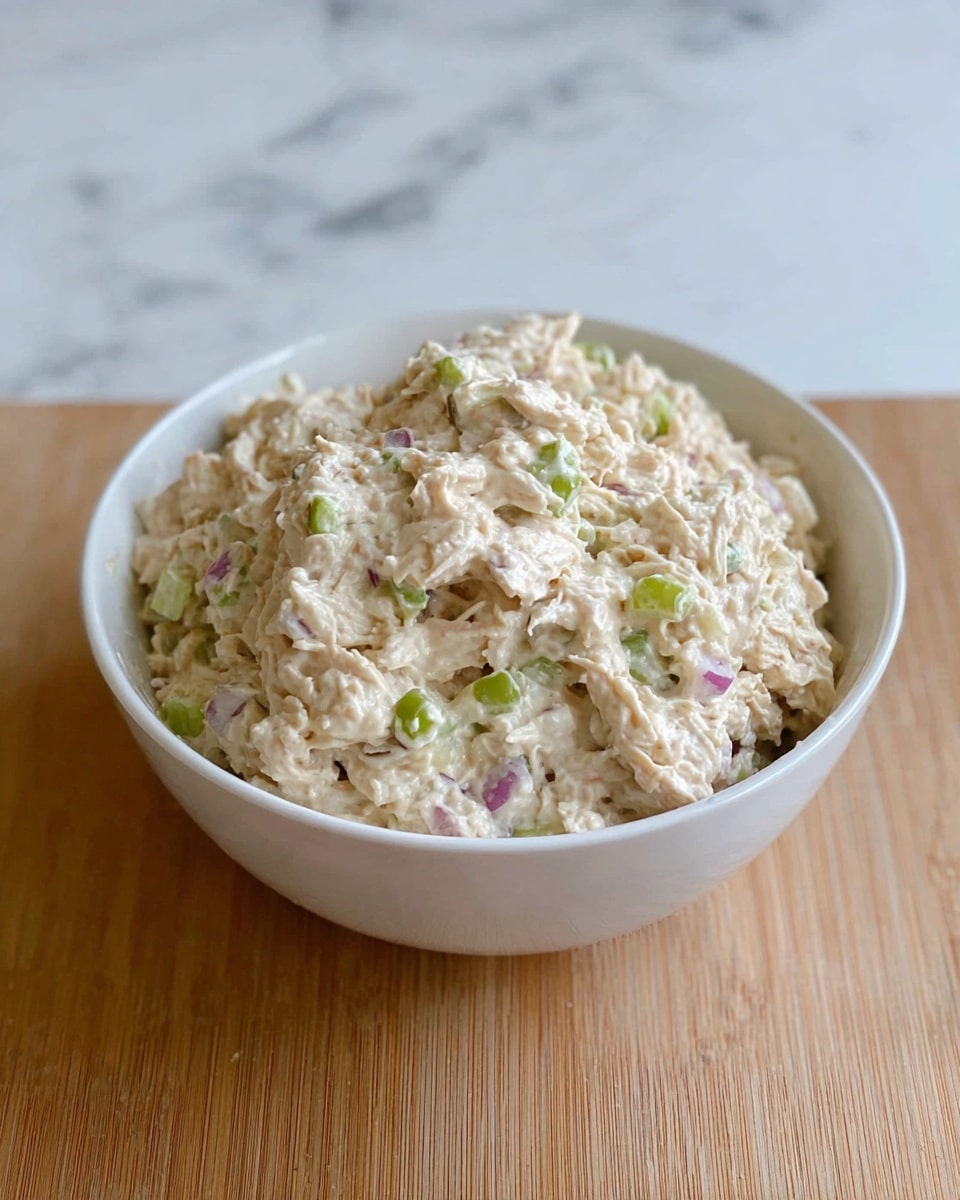 A white bowl filled with a thick, creamy mixture that looks like chicken salad. The salad has a pale beige color with small green celery pieces and tiny bits of purple onion scattered throughout. The texture of the salad is chunky and slightly fluffy, showing the shredded chicken mixed evenly with the creamy dressing. The bowl sits on a white marbled surface. photo taken with an iphone --ar 4:5 --v 7