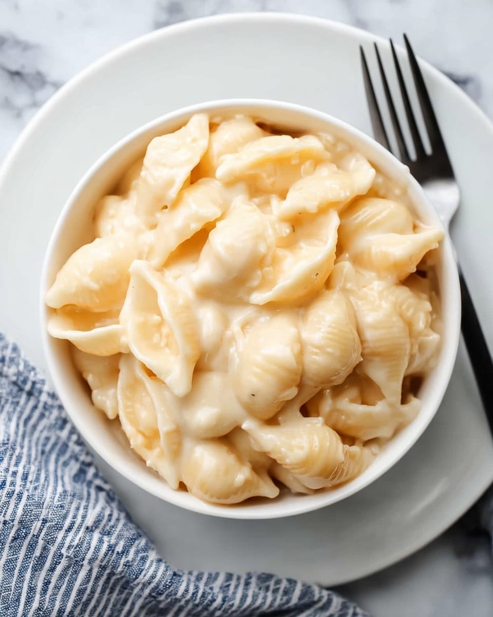 The image shows a white round bowl filled with creamy macaroni and cheese made with shell pasta. The pasta shells are fully covered in a thick, smooth cheese sauce that is pale yellow and looks rich and gooey. The bowl sits on a matching white plate, placed on a surface with a white marbled texture. To the right of the bowl, there is a fork with a black handle resting on the edge of the plate. A blue and white striped cloth is folded and placed near the fork. The photo is taken from above, focusing tightly on the bowl and the creamy dish inside. photo taken with an iphone --ar 4:5 --v 7