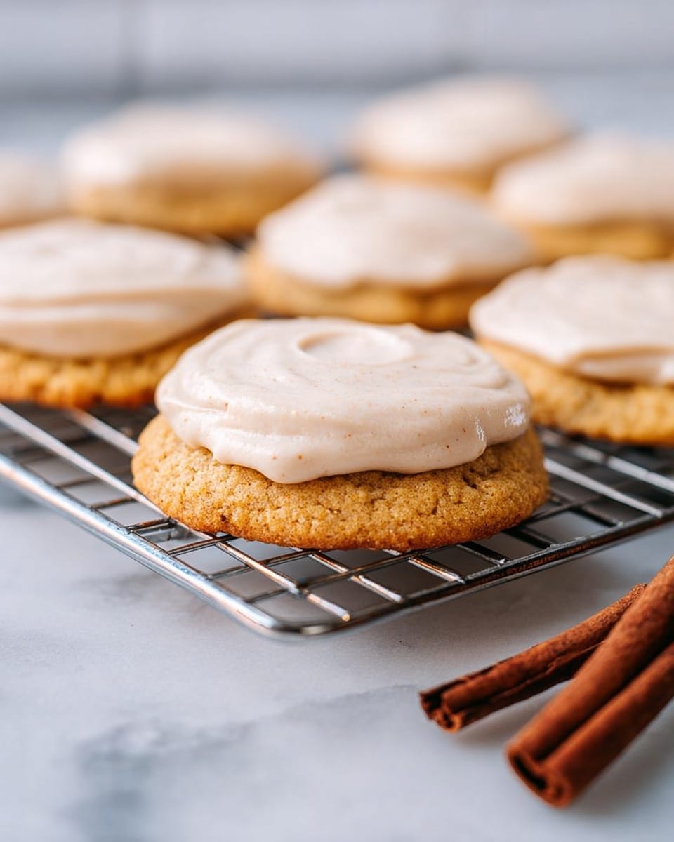 The image shows soft, round cookies on a metal cooling rack placed on a white marbled texture surface. Each cookie has two layers: the base is a golden brown textured cookie, and the top is a creamy, light beige frosting layer spread smoothly but with soft, wavy patterns. In the foreground, two cinnamon sticks with a rich brown color and rough texture lay on the surface, adding a cozy touch. The background is softly blurred, highlighting the cookies in the front. Photo taken with an iphone --ar 4:5 --v 7