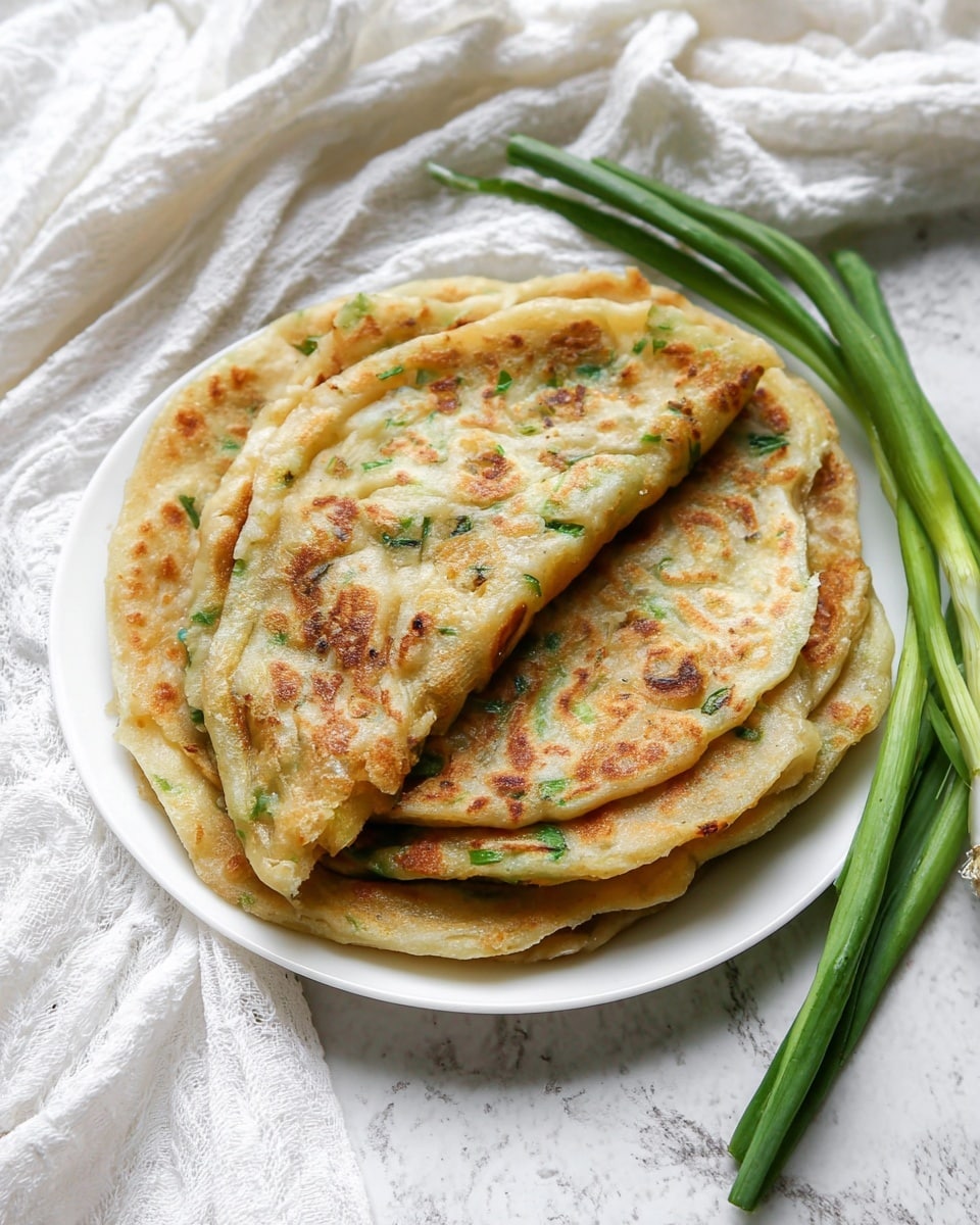A white plate with a stack of four round, golden-brown scallion pancakes showing crispy, flaky textures and small green scallion pieces throughout. The top pancake is partially folded to reveal its soft, layered inside. Three fresh green scallions lie on the right side of the plate. The background is a white marbled texture with soft white cloth draped around. photo taken with an iphone --ar 4:5 --v 7