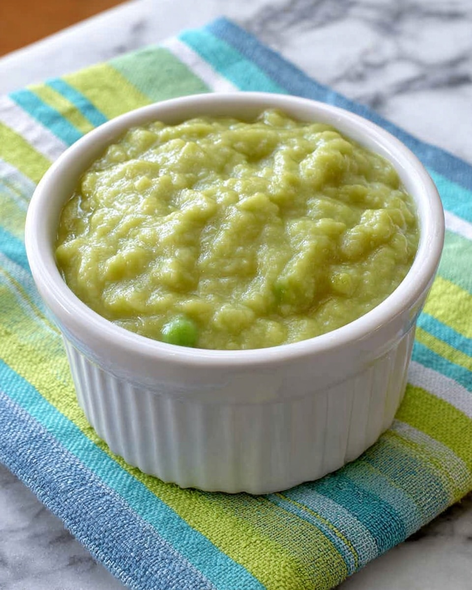 A white ceramic ramekin filled with chunky green pea mash that has a thick, slightly lumpy texture with some visible pea pieces. The ramekin sits on a colorful woven placemat with stripes in bright green, blue, aqua, and white, placed on a white marbled surface. A silver spoon is partially visible on the left side near the ramekin. The lighting is natural, showing the smooth and coarse parts of the mash clearly. photo taken with an iphone --ar 4:5 --v 7