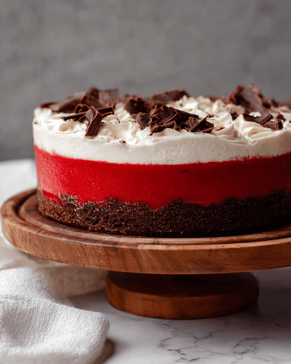 The image shows a round layered dessert with three clear layers placed on a wooden stand. The bottom layer is dark brown and looks like a crunchy or crumbly crust. The middle layer is thick and bright red, smooth and shiny in texture. The top layer is a thick spread of soft, white cream with some rough folds and swirls. Small dark chocolate shavings are scattered on top of the cream. The background has a simple gray color, and the surface beneath the stand has a white marbled texture with a white cloth partly visible near the bottom. photo taken with an iphone --ar 4:5 --v 7