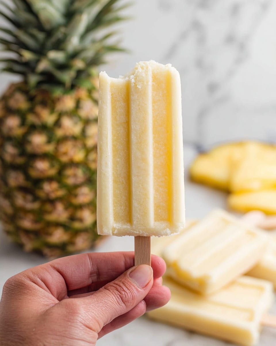 A close-up image shows a woman's hand holding a light yellow popsicle with a bite taken from the top right corner. The popsicle has vertical ridges and a soft, icy texture. In the background, there is a whole pineapple with green and yellow skin and several more popsicles lying on a white marbled surface. The overall scene looks fresh and cool. photo taken with an iphone --ar 4:5 --v 7