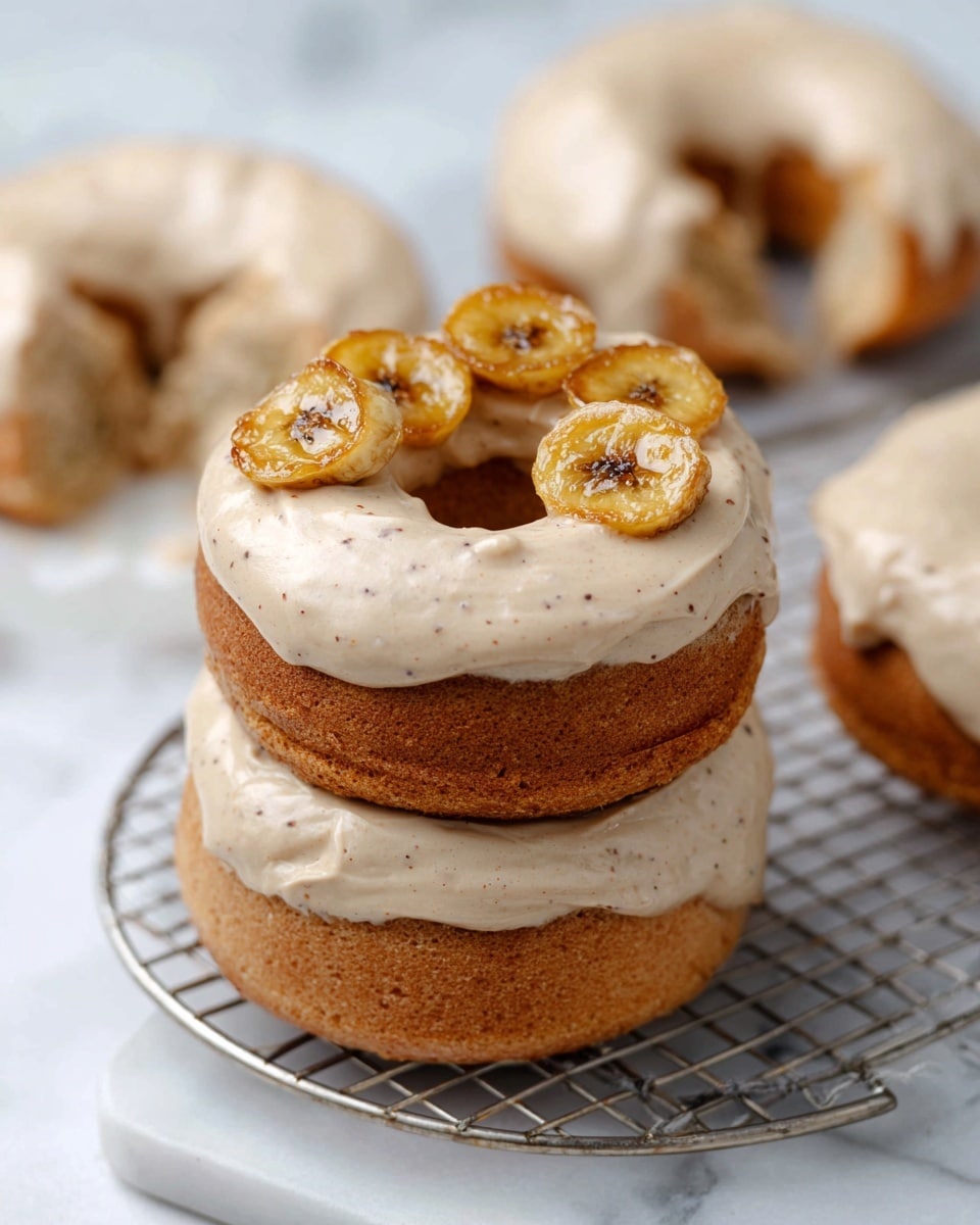 A stack of three donut-shaped cakes sits on a silver wire rack above a white marbled surface, each cake looking light brown with a soft texture. Each cake is covered with a thick layer of creamy, light beige frosting that has tiny brown specks mixed in, evenly spread over the top. On the top cake, three small round slices of golden-brown caramelized banana rest side by side, adding a shiny, slightly crispy texture. In the blurry background, two more cakes with the same frosting are visible, one with a bite taken out of it. Photo taken with an iphone --ar 4:5 --v 7