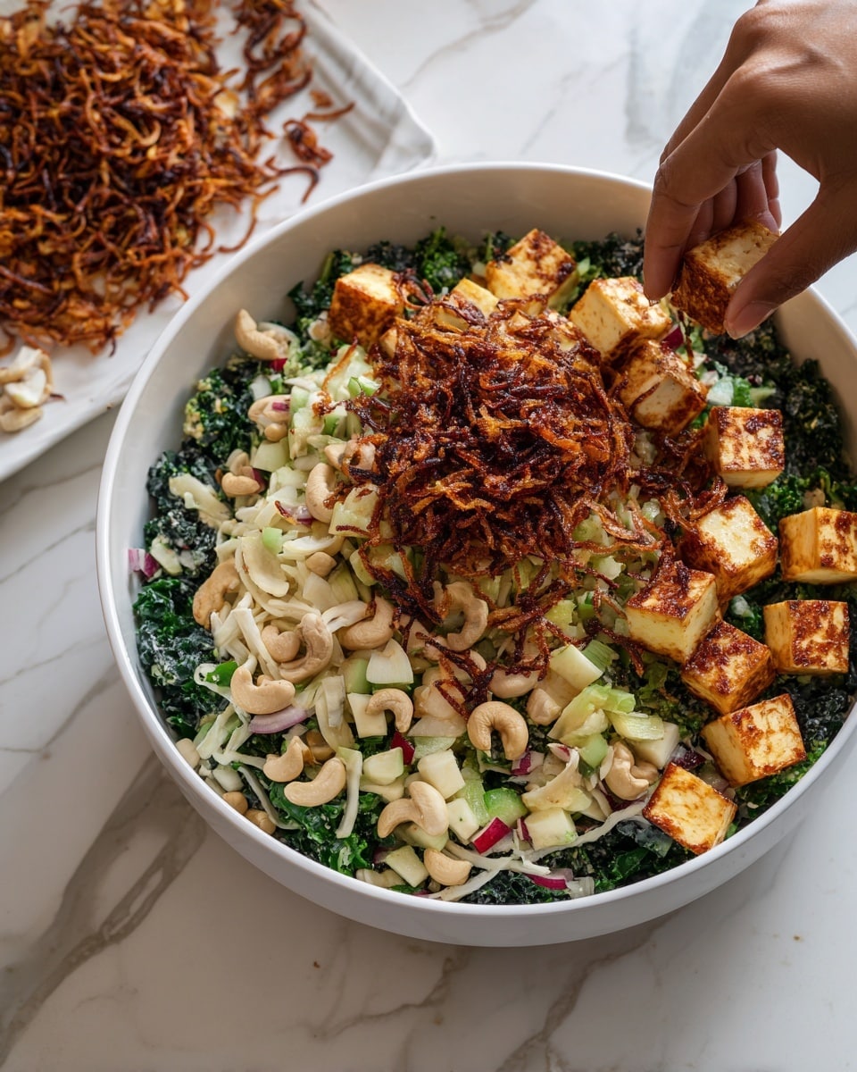 A white bowl contains a layered salad starting with a bottom layer of dark green shredded kale. Above that is a layer of chopped cashew nuts in pale beige color, followed by a layer of white and red chunks of apple pieces and green chopped celery. On top are golden brown grilled paneer cubes. The final top layer is sprinkled with crispy fried onions in reddish-brown color. A woman's hand is adding some of the crispy fried onions to the salad. To the left of the bowl is a white plate with more crispy fried onions. The background surface is white with a marbled texture. photo taken with an iphone --ar 4:5 --v 7