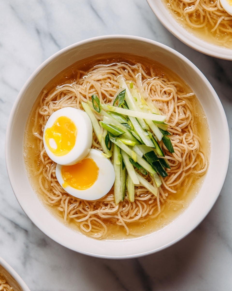 A stainless steel bowl filled with clear golden broth holding three layers: at the base, thin brown noodles twisted in a nest shape; on top of the noodles, several thin slices of light pink cooked meat; and on the meat, a halved boiled egg with a bright yellow yolk, surrounded by thin strips of fresh green cucumber and white radish. The bowl is placed on a white marbled surface. Photo taken with an iphone --ar 4:5 --v 7