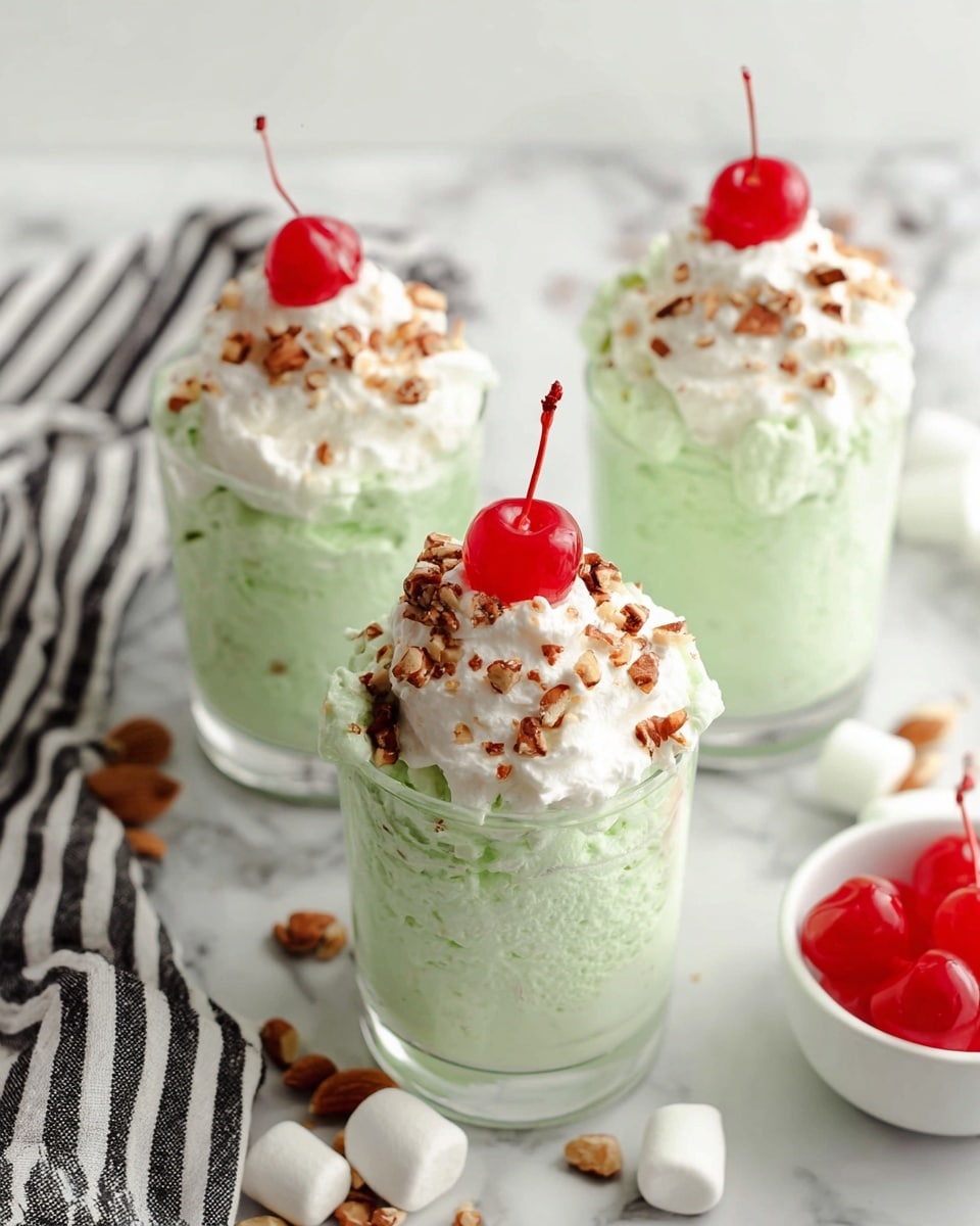 Three clear glasses each hold a fluffy, light green mousse with a creamy texture, showing bits of small nuts mixed inside. Each glass is topped with a small dollop of white whipped cream, sprinkled with chopped brown nuts, and crowned with a bright red cherry with a stem. The glasses are placed on a white marbled surface, with a striped black and white cloth nearby, along with some scattered small white marshmallows and chopped nuts. A white bowl with extra cherries is partly visible on the side. Photo taken with an iphone --ar 4:5 --v 7