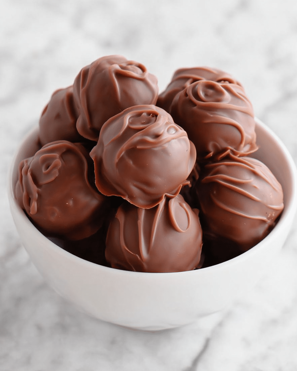 A white bowl is filled with round chocolate treats that have a swirled, uneven surface texture, showing thick layers of milk chocolate coating. One of the treats is cut in half and placed on top, revealing three inner layers: a light beige cookie layer on top and bottom, a dark brown chocolate layer in the middle, and a creamy peanut butter layer above the bottom cookie, all layered inside the milk chocolate shell. The bowl rests on a white marbled surface, and a blurred orange bag of Reese's minis is seen in the background. photo taken with an iphone --ar 4:5 --v 7