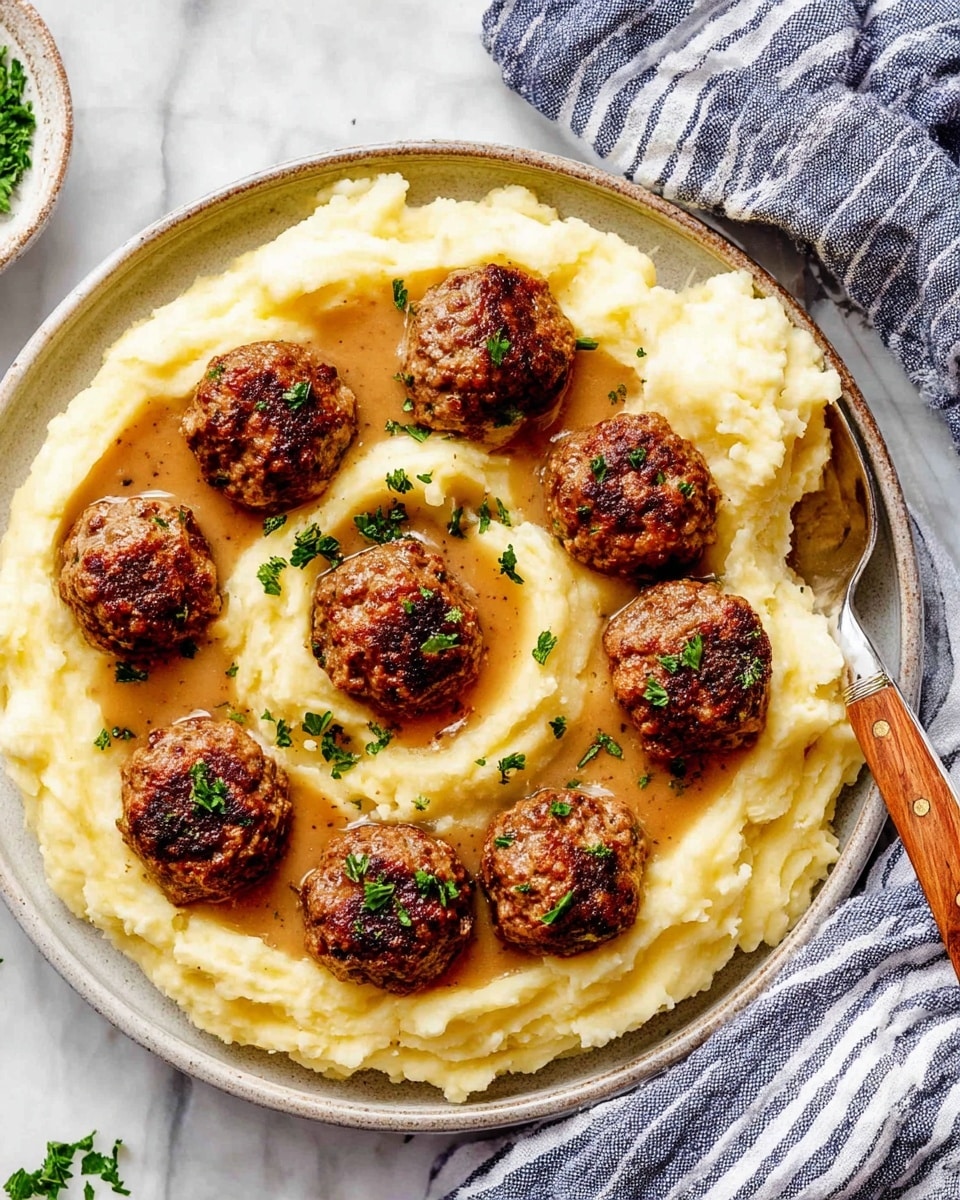 A white round plate holds a base layer of creamy, pale yellow mashed potatoes spread in a thick, uneven layer with soft ridges and swirls. On top of this is a layer of rich, light brown gravy partially covering the center. The top layer consists of ten evenly spaced, browned meatballs with a textured surface, each garnished with small green parsley leaves. A spoon with a wooden handle rests on the side of the plate, partly in the mashed potatoes and gravy. The plate is placed on a white marbled surface with a blue and white striped cloth partially visible nearby. Photo taken with an iphone --ar 4:5 --v 7