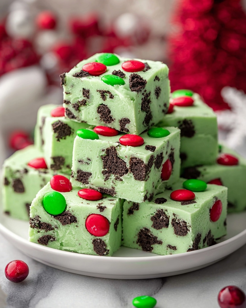 A stack of seven square pieces of green fudge is shown on a white plate, each piece mixed with black cookie chunks inside and topped with red and green candy-coated chocolates. The fudge has a smooth, creamy texture with unevenly placed chocolate cookie bits embedded throughout each layer. The candies on the top give a festive look with their bright glossy colors. In the background, there are blurred red berry decorations and a white marbled surface. photo taken with an iphone --ar 4:5 --v 7