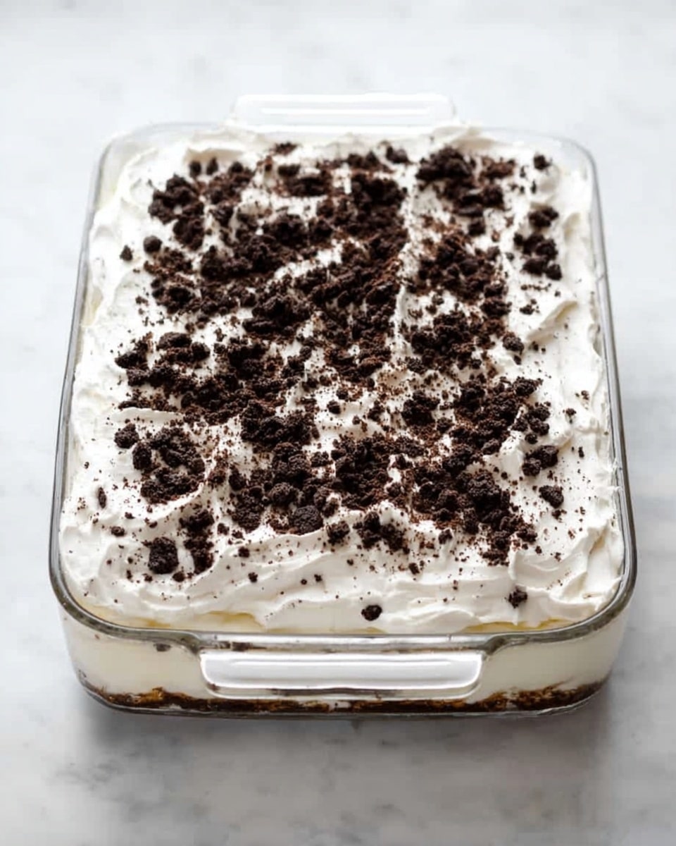 A clear glass rectangular baking dish with two handles contains a dessert with two visible layers. The bottom layer is creamy white and smooth, filling most of the dish. On top is a thick layer of white whipped cream, spread evenly but with soft peaks. The whipped cream is covered with a generous amount of dark brown crumbled cookies scattered unevenly across the surface, creating a rough texture and contrast against the white cream. The dish is placed on a white marbled texture surface. photo taken with an iphone --ar 4:5 --v 7