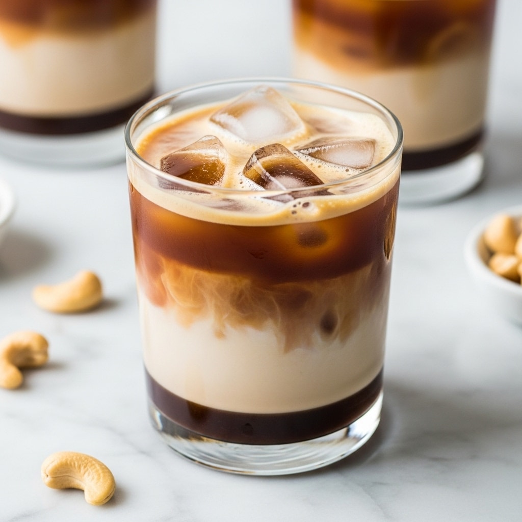 A clear glass shows a drink with three layers: the bottom layer is a dark coffee color with a rich, thick texture; above it is a creamy light beige layer blending softly with the coffee; the top layer is frothy white with ice cubes floating inside. The glass sits on a white marbled surface with others in the background, along with some scattered cashew nuts adding a relaxed feel. photo taken with an iphone --ar 4:5 --v 7