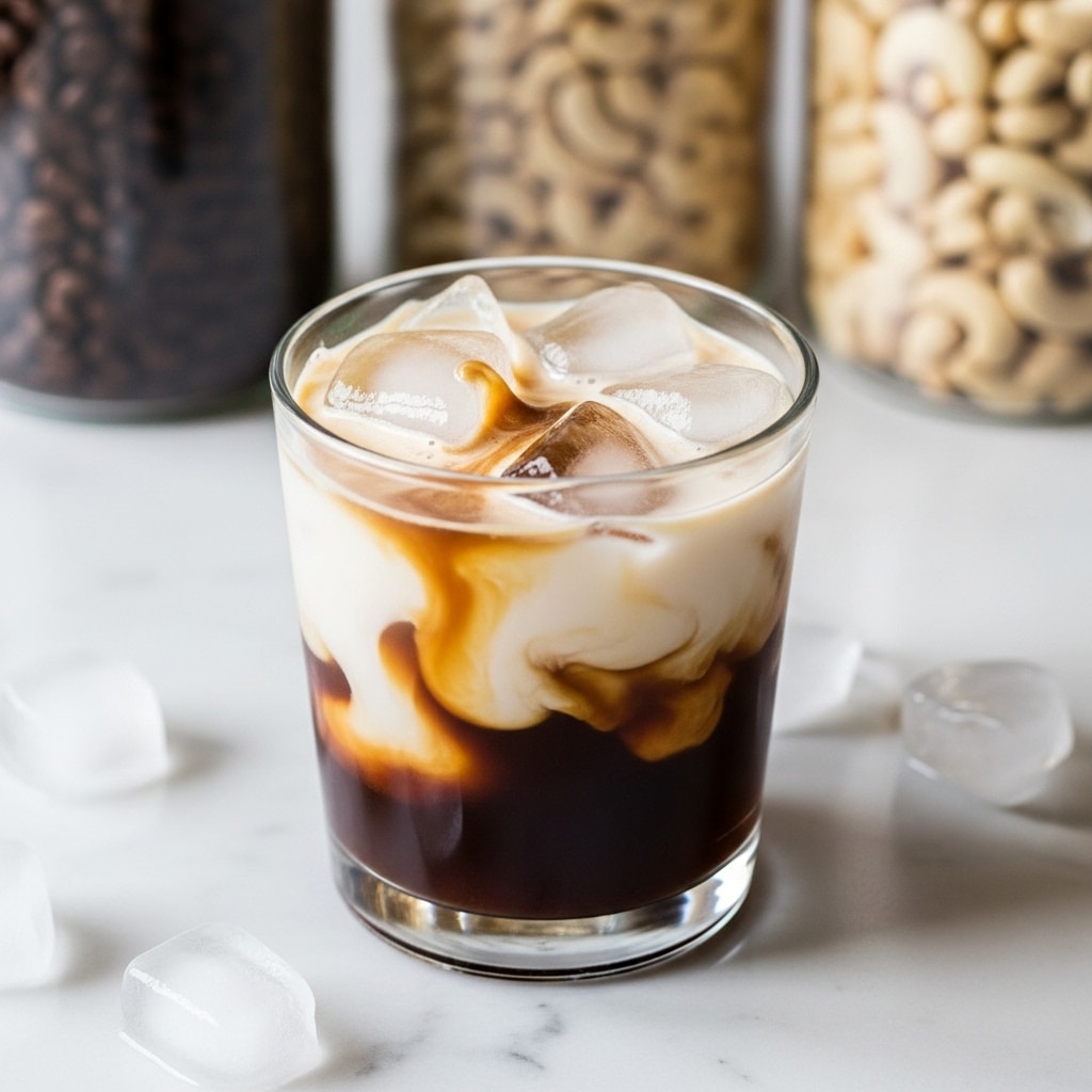 A clear glass holds a layered drink on a white marbled surface. The bottom layer is dark brown, thick coffee or ale, with a creamy white layer above it that swirls softly as it meets the ice cubes floating on top. The ice cubes are transparent and scattered unevenly, with light reflecting off their surfaces. In the blurred background are glass jars filled with coffee beans and white cashews, adding texture but keeping focus on the drink. The overall scene is simple and fresh, showing the drink in soft natural light. photo taken with an iphone --ar 4:5 --v 7