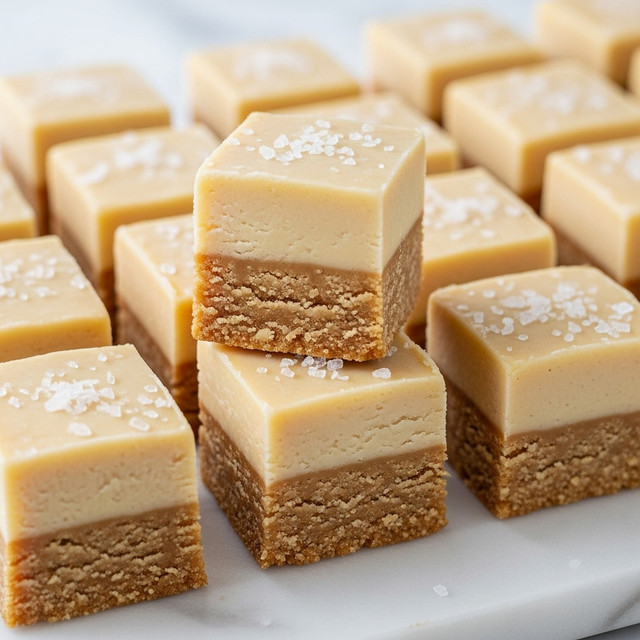 The image shows a close-up of pale beige fudge squares arranged neatly in rows on a white marbled surface. Each square has two layers: a soft, crumbly bottom layer with a slightly rough texture and a smooth, firm top layer with a pale, creamy color. Some of the fudge pieces are sprinkled with flakes of coarse salt, adding a contrast in texture. One piece is slightly raised above the others, showing the thickness and texture of both layers. The overall look is creamy and soft with a hint of salt on top. photo taken with an iphone --ar 4:5 --v 7