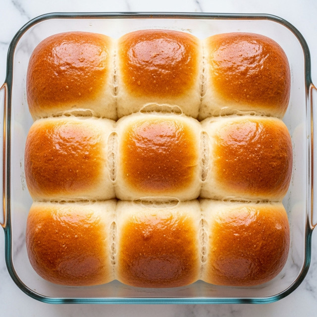 The image shows a clear glass baking dish filled with nine soft, shiny golden-brown dinner rolls, arranged in a 3x3 grid. Each roll has a smooth, round top with a slightly glossy finish, indicating they were brushed with butter or egg wash. The texture looks fluffy and light, with a few small wrinkles on the tops. The dish sits on a white marbled texture. photo taken with an iphone --ar 4:5 --v 7
