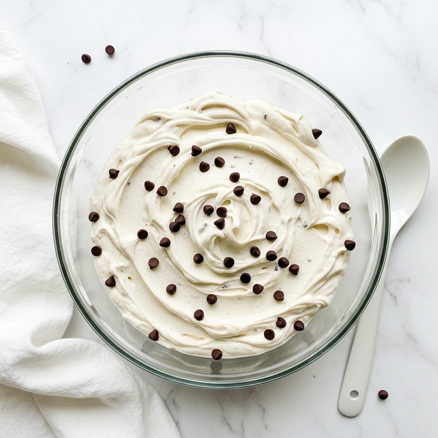 A glass bowl filled with a creamy, thick white mixture that has small dark chocolate chips spread evenly throughout. The texture looks smooth with slight swirls and peaks on the surface of the cream. The bowl sits on a white marbled texture with a white cloth napkin and a white spoon nearby. The overall look is fresh and inviting. photo taken with an iphone --ar 4:5 --v 7
