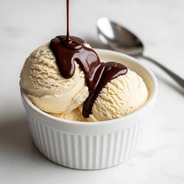 A small white bowl with vertical ridges holds two big scoops of light cream-colored ice cream with a soft, slightly rough texture. Dark, glossy chocolate sauce is being poured over the top, creating thick layers that slowly flow down the ice cream, adding a rich contrast to the pale scoops. The bowl sits on a smooth white marbled surface with a silver spoon placed behind it. Photo taken with an iphone --ar 4:5 --v 7