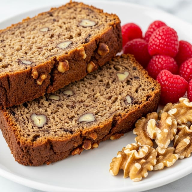 Two thick slices of moist banana nut bread with a light brown color and dark specks of banana and nut pieces are stacked slightly leaning on each other on a white plate. The texture looks soft and crumbly with visible small nuts and banana bits throughout the bread. On the right side of the plate, there are several bright red raspberries and a small pile of walnut halves resting on the white marbled surface. The close-up view highlights the bread’s dense, slightly grainy texture and fresh ingredients. Photo taken with an iphone --ar 4:5 --v 7