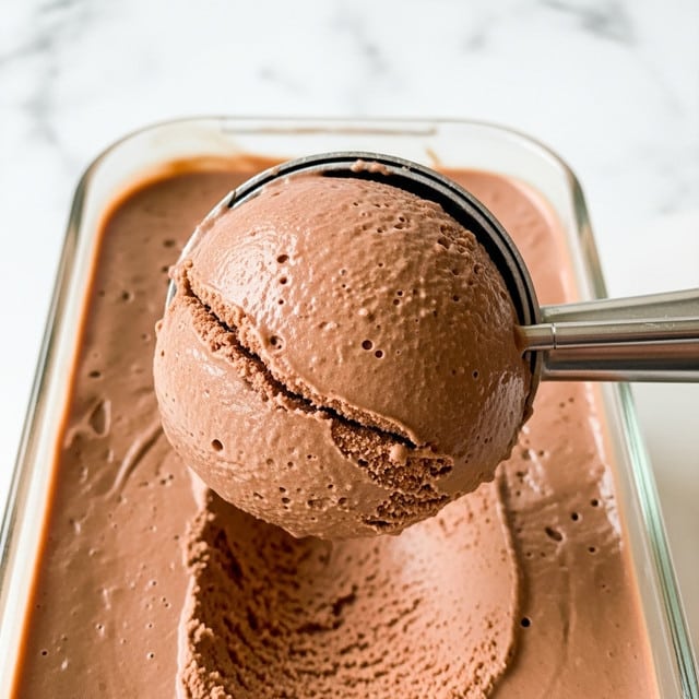 The image shows a close-up of two scoops of smooth and creamy chocolate ice cream in a white bowl with a dark rim. The top scoop is more rounded with a velvety surface while the bottom scoop has a textured, slightly rough surface that reveals the ice cream’s creaminess. A shiny metal spoon is placed inside the bowl, leaning against the ice cream. The bowl sits on a white marbled surface. photo taken with an iphone --ar 4:5 --v 7
