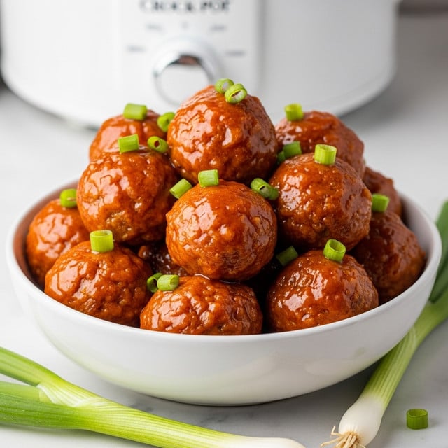 A white bowl filled with about three layers of round meatballs coated in a shiny, bright orange sauce, each meatball showing a slightly textured surface beneath the glaze. The top layer of meatballs is garnished with small, chopped green onion pieces scattered unevenly. The bowl rests on a white marbled surface, with some whole green onions placed in the foreground. A white slow cooker with a visible temperature dial is in the blurred background, adding context to the dish. Photo taken with an iphone --ar 4:5 --v 7