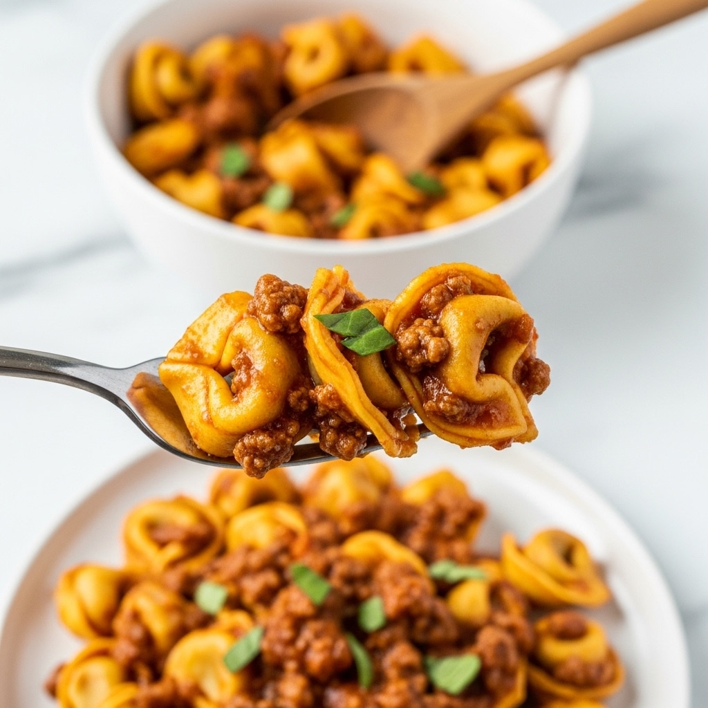 A close-up view of a silver fork holding three pieces of tortellini pasta coated in a rich, thick, reddish-brown meat sauce with bits of ground meat and small green herb leaves sprinkled on top. In the background, there is a white bowl filled with more tortellini mixed with the same sauce, along with a wooden spoon resting inside it. Below, a white plate full of tortellini pasta covered in the meaty sauce sits on a white marbled surface. The focus is sharp on the fork with the pasta, while the background is softly blurred. photo taken with an iphone --ar 4:5 --v 7