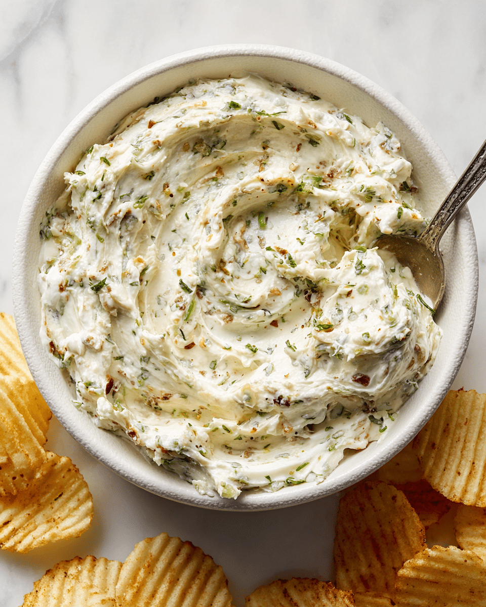A close-up view of a bowl filled with a creamy dip that has a thick, slightly chunky texture showing bits of herbs and ingredients mixed throughout. The dip is off-white with small green and brown specks, swirled smoothly on top with some peaks and folds, with a silver spoon partially submerged in the dip on the right side. The bowl is white with a slightly rustic rough texture visible on the inside rim. Around the bowl, several ridged potato chips are scattered on a white marbled surface. Photo taken with an iphone --ar 4:5 --v 7