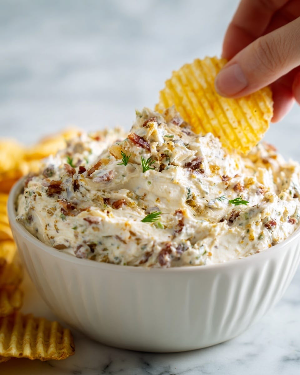 A close-up view of a creamy dip shown in a white bowl with soft, thick texture that has visible small green and light brown pieces mixed throughout. A yellow ridged potato chip is being dipped into the thick cream, held by a woman's hand on the right side of the image. The dip looks rich and smooth with a slightly shiny surface and a layered mix of colors mainly off-white with specks of green and brown, all set on a white marbled textured background. photo taken with an iphone --ar 4:5 --v 7