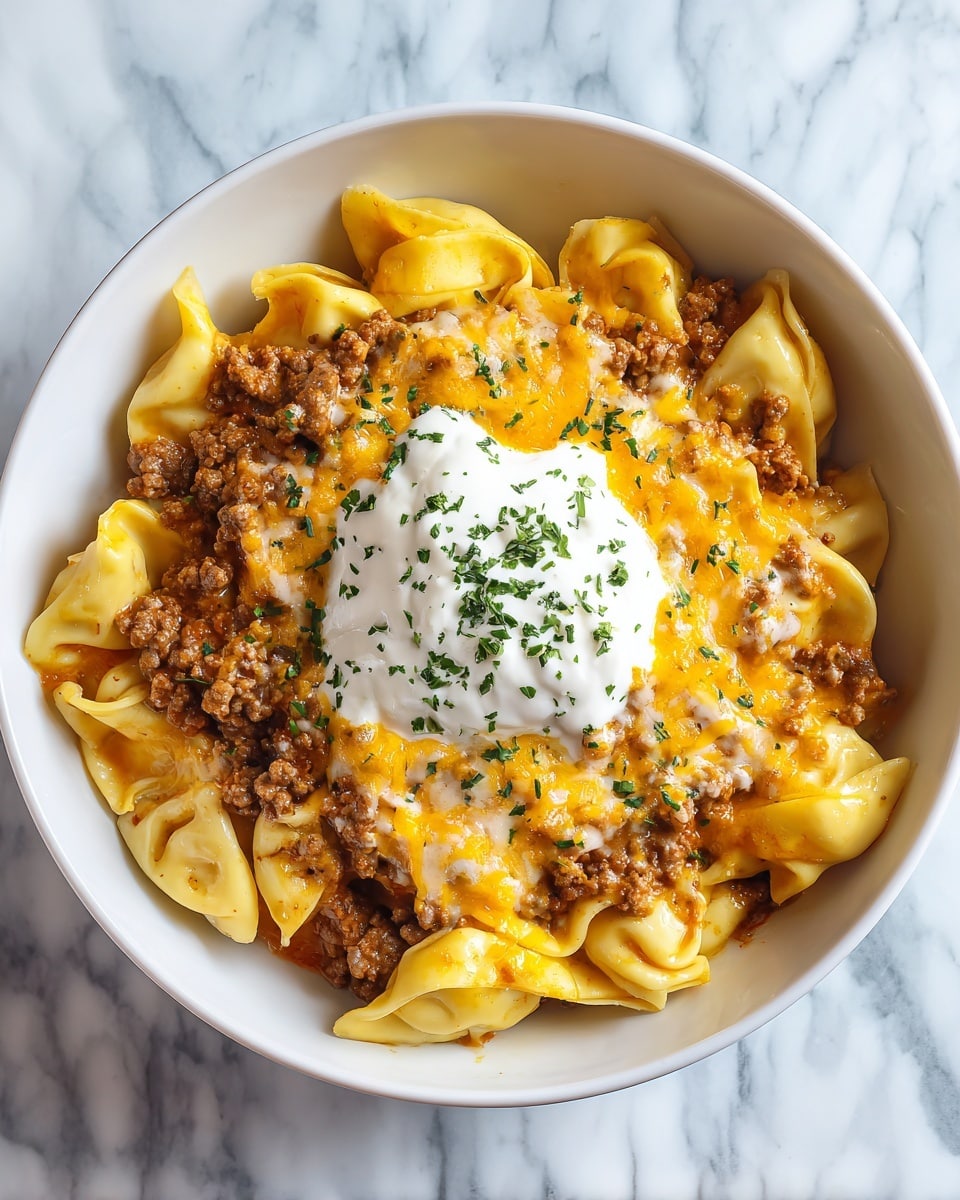 A white bowl filled with creamy tortellini pasta mixed with small pieces of ground meat, topped with a dollop of white sour cream sprinkled with green herbs, and shredded orange cheddar cheese. At the back edge of the bowl rests a golden-brown crispy piece of toast. The creamy sauce coats the pasta, which has small green flecks inside, and the dish is set on a white marbled texture. photo taken with an iphone --ar 4:5 --v 7
