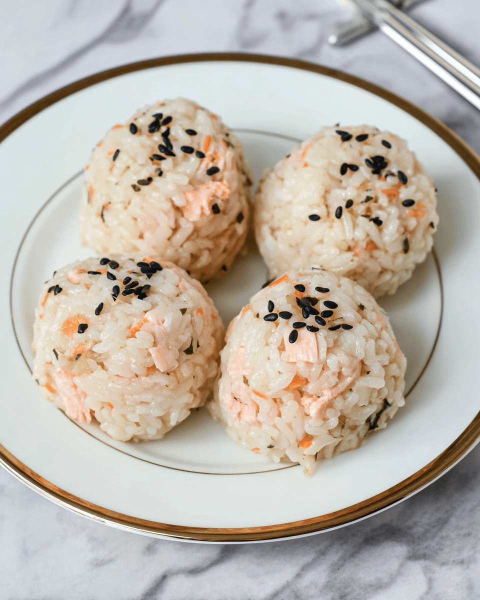 Four rice balls sit closely together on a white plate with a thin gold rim. Each ball is made of sticky white rice mixed with light pink pieces of salmon and sprinkled on top with small black sesame seeds. The rice looks soft and slightly shiny, holding its round shape well. The plate is placed on a white marbled surface, and a pair of silver chopsticks rests nearby. photo taken with an iphone --ar 4:5 --v 7