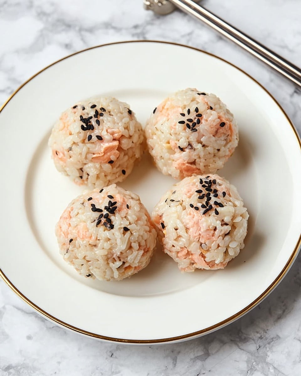 Four round rice balls sit on a white plate with a gold rim, each ball mixed with small, uneven pieces of pink salmon and topped with black sesame seeds. The rice looks soft and slightly shiny, tightly packed into smooth spheres. The plate rests on a white marbled surface with a pair of silver chopsticks placed diagonally near the top right corner. The photo taken with an iphone --ar 4:5 --v 7