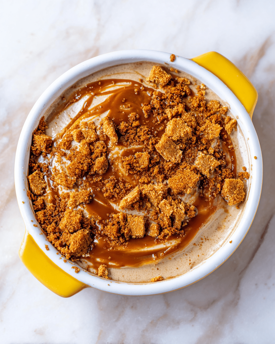 A round white ceramic dish with two small handles, covered on the outside in bright yellow, holds a layered dessert placed on a white marbled surface. The dessert has a base layer of smooth, light brown mousse or cream with a soft swirl pattern. On top, there are glossy caramel or toffee drizzles spread unevenly, creating glistening patches. Crumbled golden brown biscuit pieces are scattered heavily over the caramel drizzle, adding a crunchy texture that spills slightly onto the dish edges and the surrounding surface. photo taken with an iphone --ar 4:5 --v 7