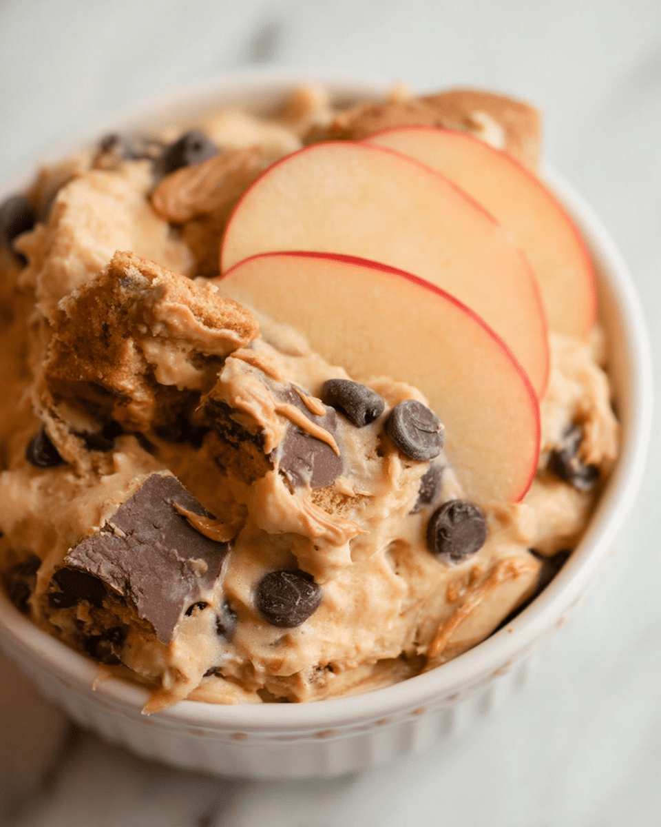 A close-up view of a white bowl filled with a creamy light tan cookie dough-like dip with visible dark chocolate chips mixed in. The top layer is decorated with swirls of smooth peanut butter and scattered chunks of chocolate peanut butter cups. The bowl sits on a white marbled surface surrounded by golden pretzels, fresh red strawberries, and light brown sandwich cookies with a cream filling. Photo taken with an iphone --ar 4:5 --v 7
