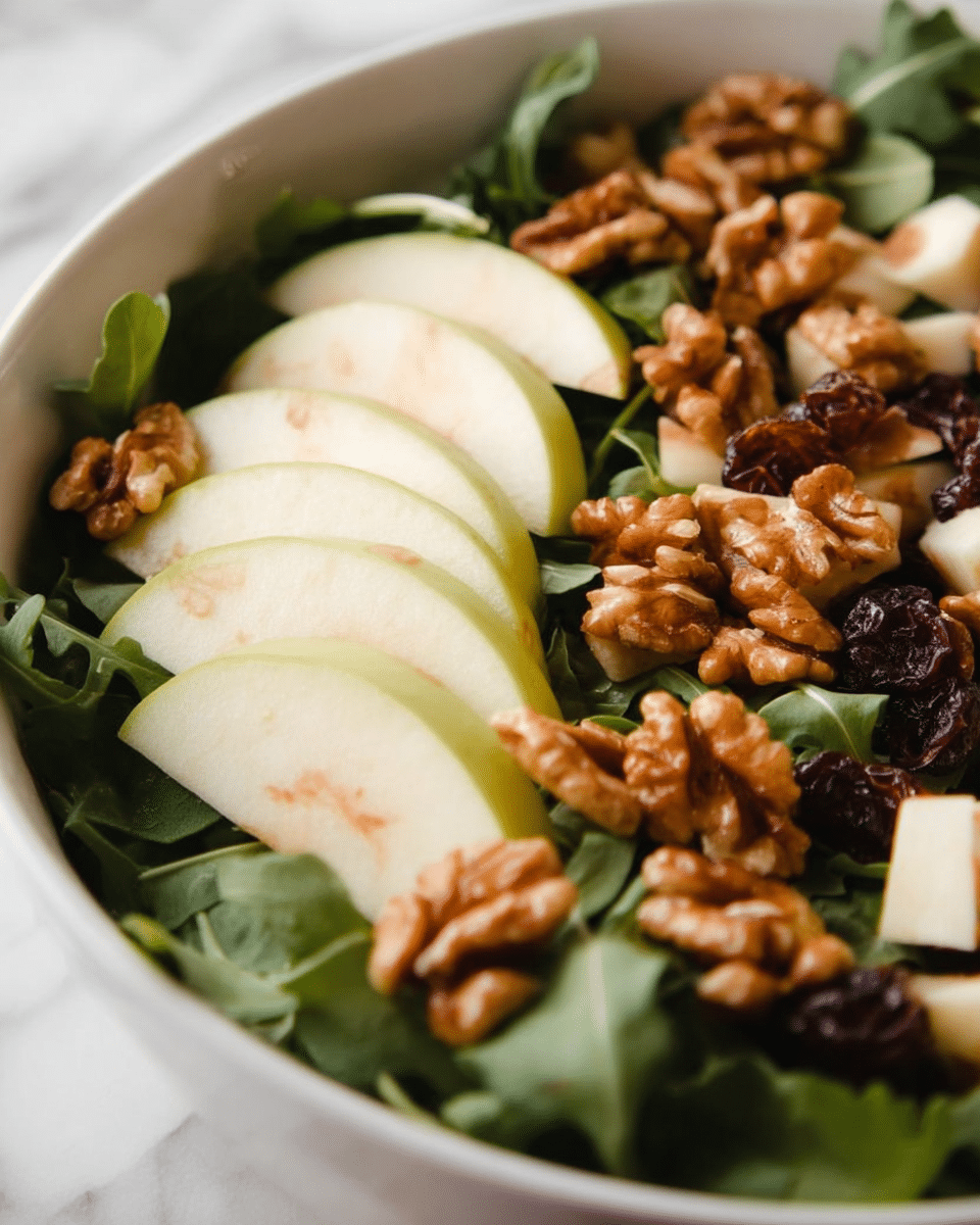 A white bowl holds a fresh salad with three main layers: the base layer is bright green leafy arugula with a slightly wrinkled texture, the middle layer contains large slices of light green apples with white flesh arranged on top, and the top layer is made of whole walnuts with a rough brown surface scattered evenly. There are also small pieces of white cheese and dark dried fruits nestled among the leaves. The background is a white marbled texture. Photo taken with an iphone --ar 4:5 --v 7
