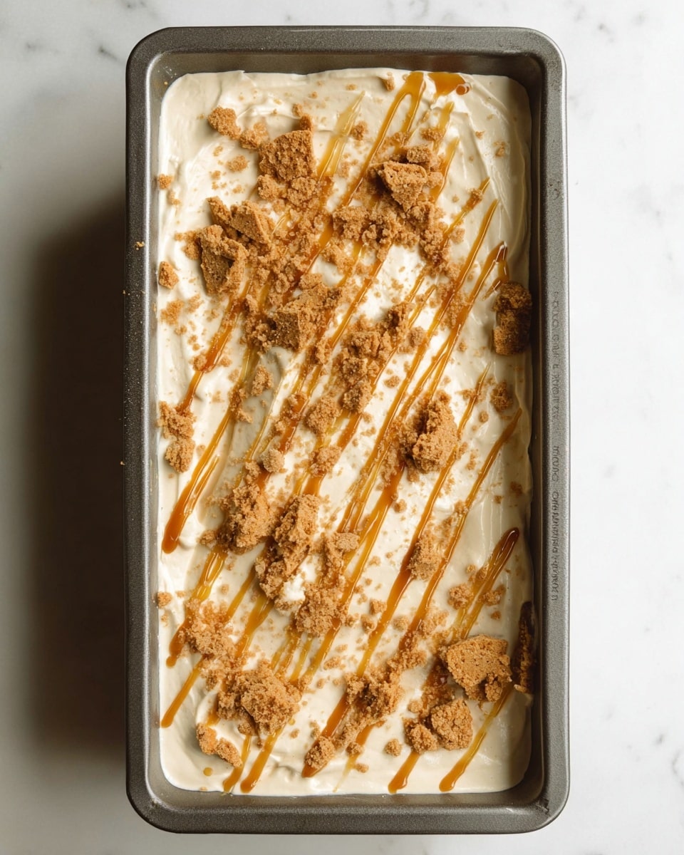 A rectangular loaf pan filled with a creamy, off-white dessert base that is smooth and slightly whipped in texture. The top is scattered with broken light brown cookie pieces, providing a rough and crumbly texture contrast. Thin caramel-colored syrup is drizzled over the entire surface in diagonal lines, adding a glossy, sticky shine. The pan sits on a white marbled surface. photo taken with an iphone --ar 4:5 --v 7