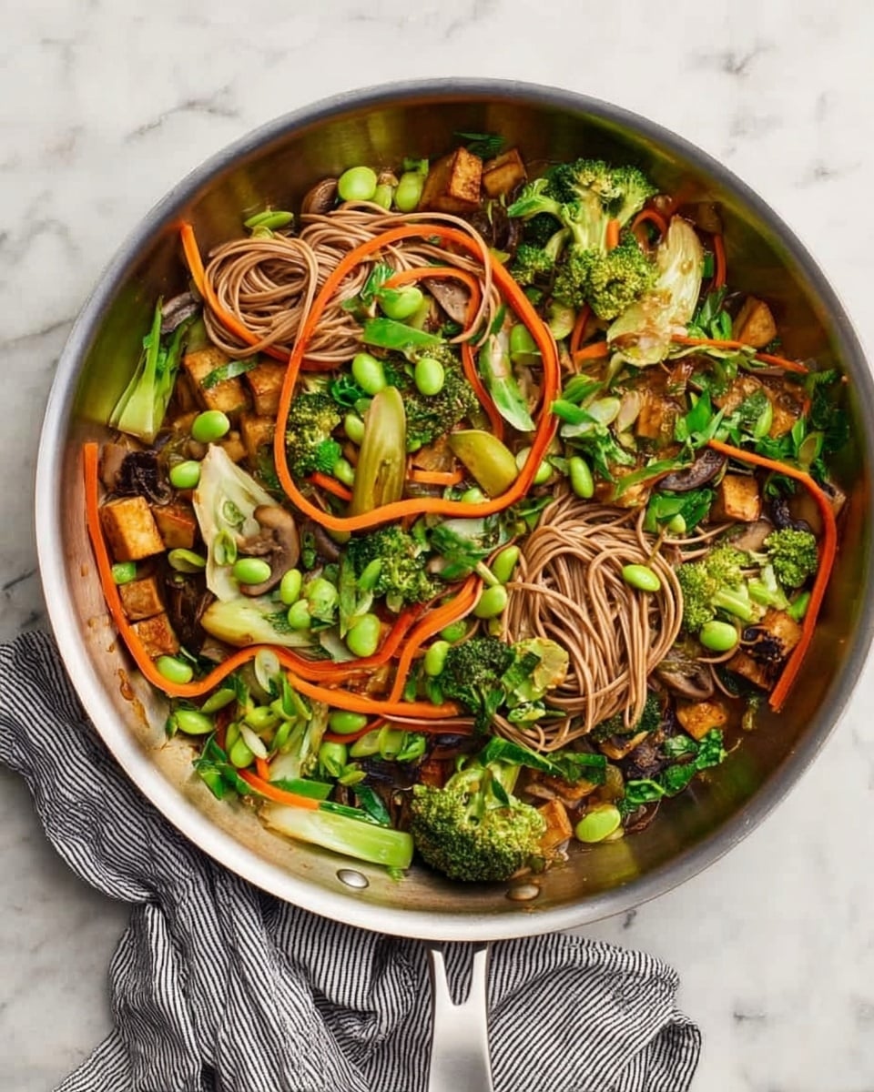 A metal pan filled with a colorful stir-fry dish sits on a white marbled surface, with a gray striped cloth nearby. The dish contains several layers: the bottom layer includes browned pieces of tofu and mushrooms with a slightly crispy texture, scattered around thick green broccoli florets and light green bok choy stems. On top of this, there are thin, soft brown noodles loosely arranged across the dish. Bright orange carrot ribbons curl gently over the noodles, while scattered vibrant green edamame beans and small leafy greens add fresh pops of color throughout. The mixture looks fresh, healthy, and vibrant with varied textures. Photo taken with an iphone --ar 4:5 --v 7