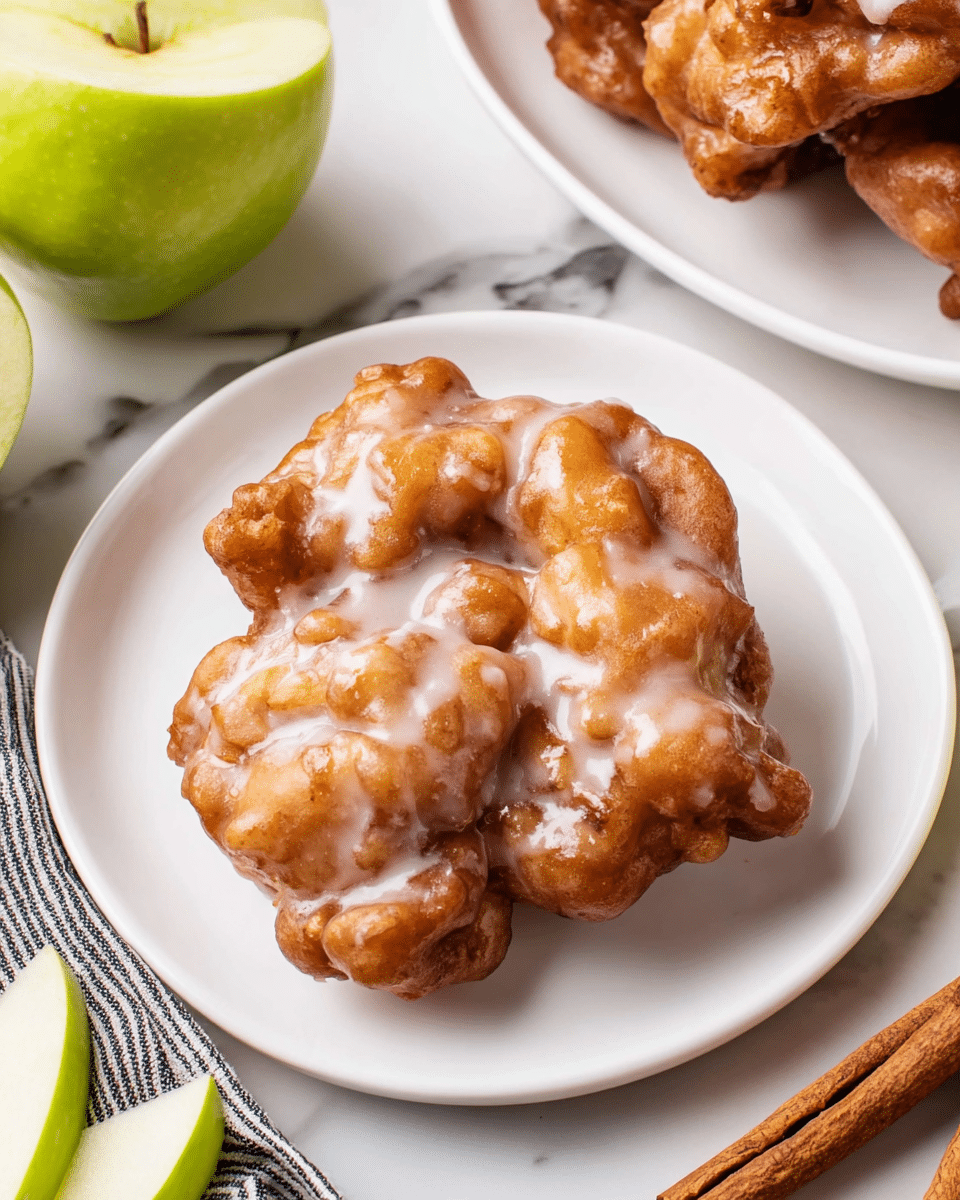 A single apple fritter sits in the center of a clean white plate, its uneven, golden-brown surface covered with a shiny, light glaze that pools slightly in the crevices, giving it a glossy texture. The fritter's lumpy shape suggests a soft and crispy mix, with small bits of apple visible under the glaze. Behind it, there is a second white plate partly shown with more apple fritters piled together, slightly out of focus. To the left of the main plate are fresh green apple slices leaning against a whole apple cut in half, displaying a bright green skin and pale inside. The scene is set on a white marbled texture, with two cinnamon sticks lying at the bottom right and a black and white striped cloth partially visible on the left. Photo taken with an iphone --ar 4:5 --v 7