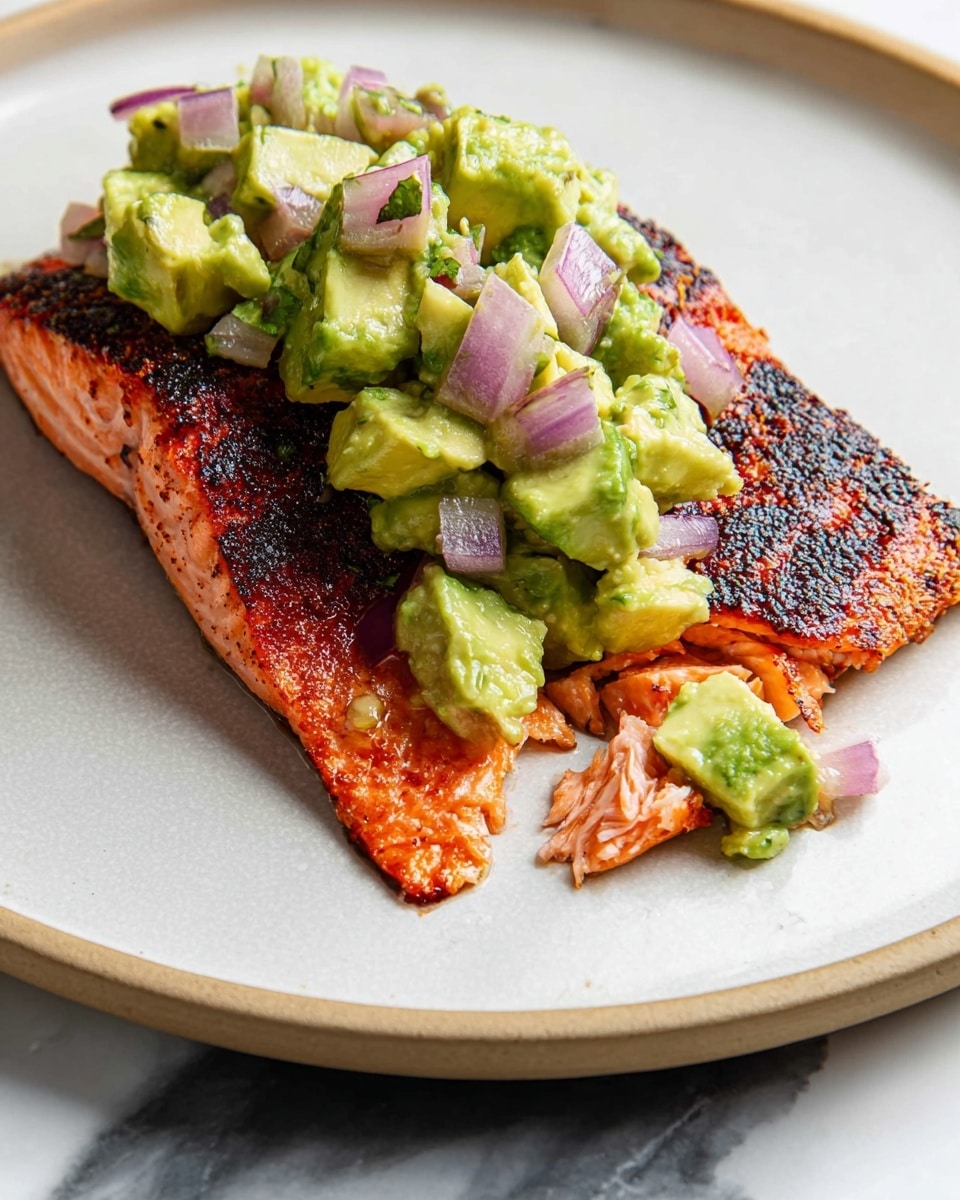 A grilled salmon fillet with a slightly charred, crispy skin on the bottom layer, showing a moist, pink interior with flaked pieces. On top, there is a chunky avocado salsa made of diced creamy green avocado and small pieces of purple-red onion, mixed together, placed in the center of the salmon. The dish is served on a smooth white plate with a simple edge, and the plate rests on a white marbled surface. photo taken with an iphone --ar 4:5 --v 7