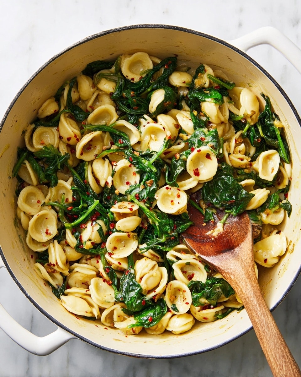 A large white pot filled with about two layers of orecchiette pasta mixed with wilted, dark green spinach leaves scattered evenly throughout. The orecchiette is pale yellow with a slightly glossy texture, and there are small red chili flakes sprinkled on top adding tiny pops of red color. A wooden spoon with a slightly browned tip rests inside the pot, partly stirring the mix. The pot is placed on a white marbled surface, enhancing the clean and simple look of the dish. Photo taken with an iphone --ar 4:5 --v 7
