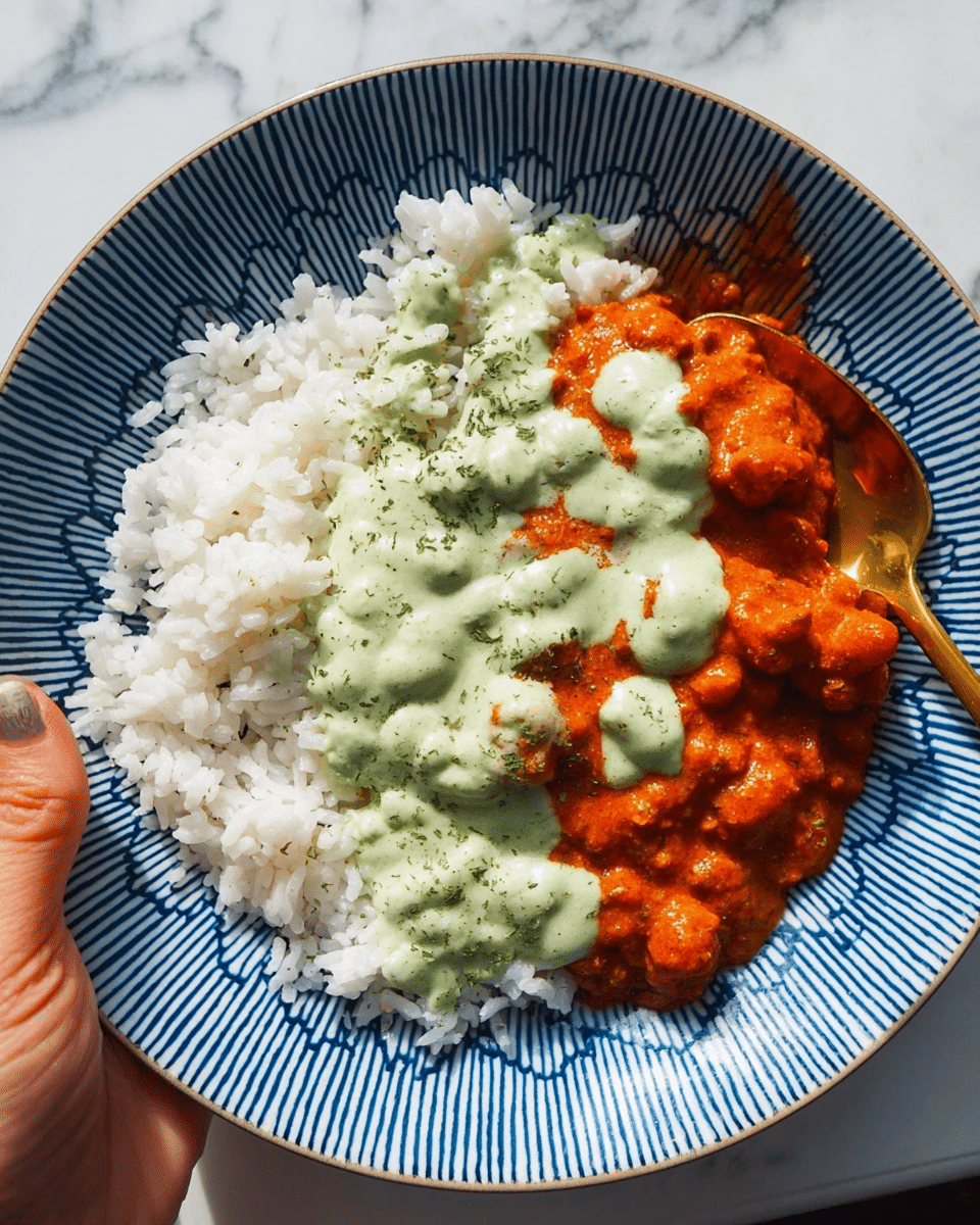 A white patterned bowl holds three main layers of food over a white marbled surface background. The bottom layer is fluffy white rice, covering about half of the bowl. On top of the rice is a thick layer of reddish-brown curry with small chickpeas scattered throughout, adding a chunky texture. A light green creamy sauce drizzles over the curry, creating a soft contrast with the warm colors. To the side, thin, curled pink pickled onions add a light, fresh touch. A small sprig of fresh parsley sits next to the onions on the rice. The bowl is held by a woman's hand wearing a blue long sleeve shirt, seen from above. Photo taken with an iphone --ar 4:5 --v 7