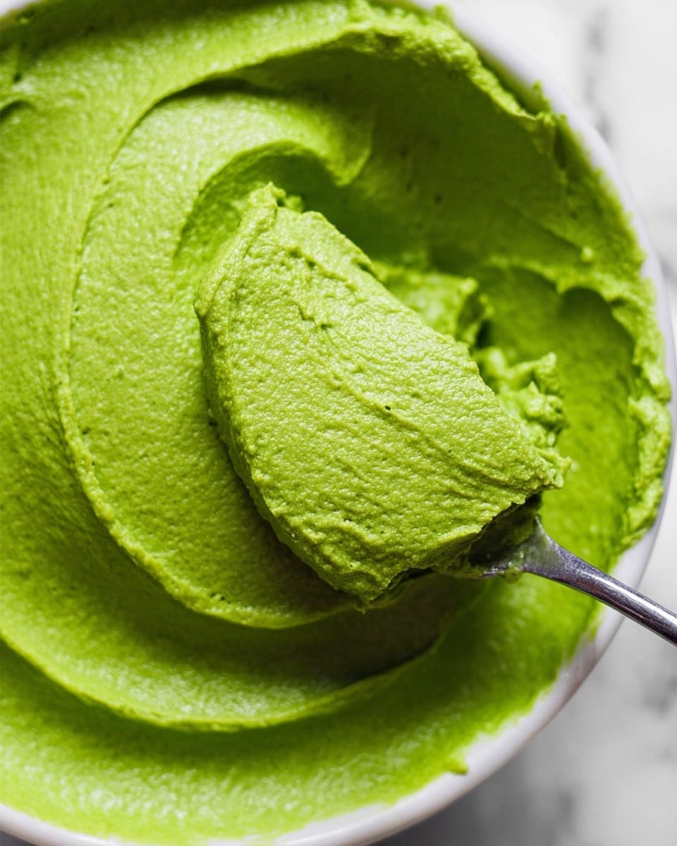 A close-up view of a smooth, thick green paste in a white bowl, showing a creamy texture with soft ridges and swirls. A silver spoon is partially dipped into the paste from the right side, lifting some of the vibrant green mixture, highlighting its dense but soft consistency. The background features a white marbled texture, adding a clean and bright contrast to the vivid green color of the paste. photo taken with an iphone --ar 4:5 --v 7