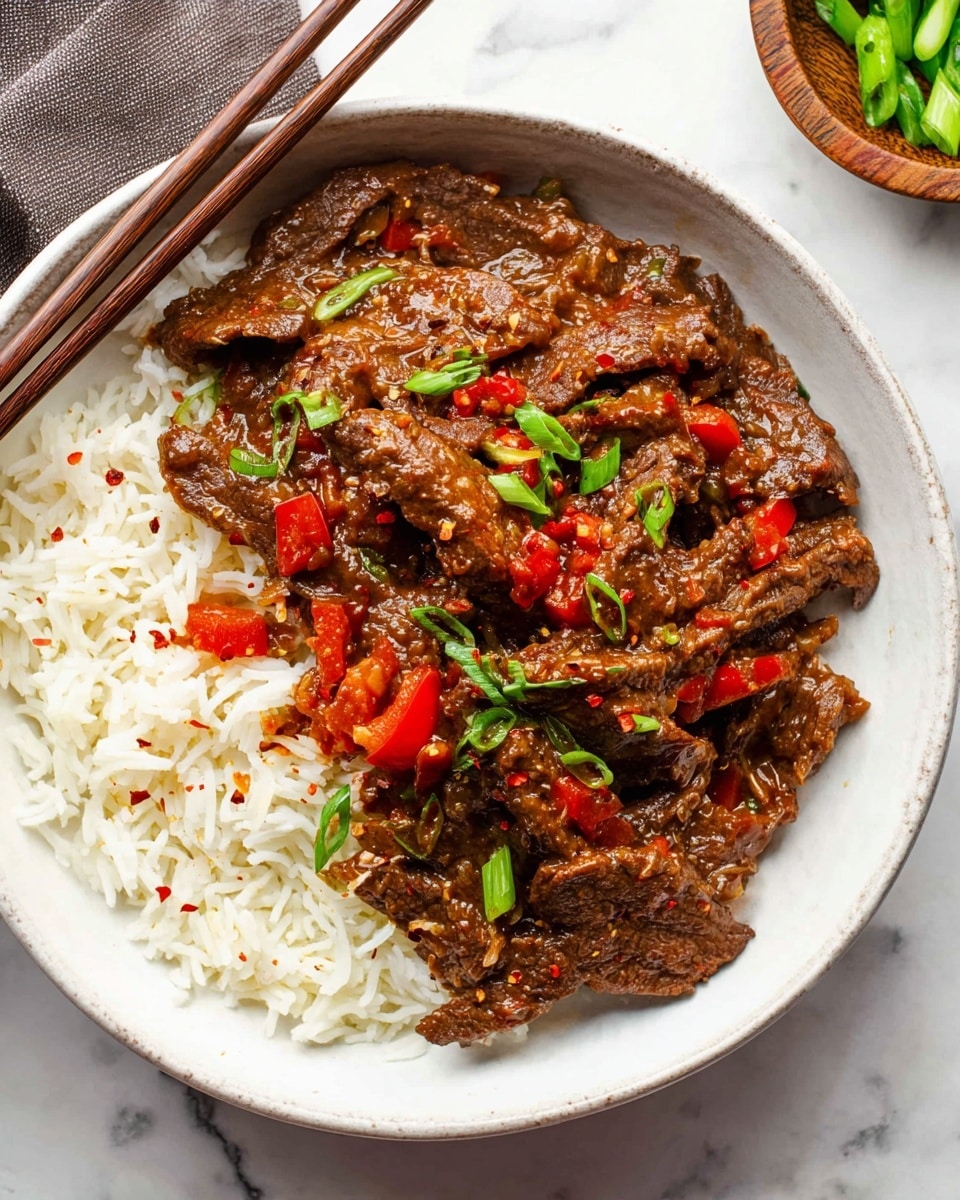 A white bowl holds a dish with two main layers: at the bottom, there is white rice with long grains, fluffy in texture; on top, there are strips of brown beef covered in a thick, shiny brown sauce mixed with small red bell pepper pieces and green onion slices scattered throughout. Some crushed red pepper flakes add specks of red on the beef. The bowl is placed on a white marbled surface, with a pair of brown chopsticks resting above it. Part of a small wooden bowl with green vegetables is seen in the top right corner. photo taken with an iphone --ar 4:5 --v 7