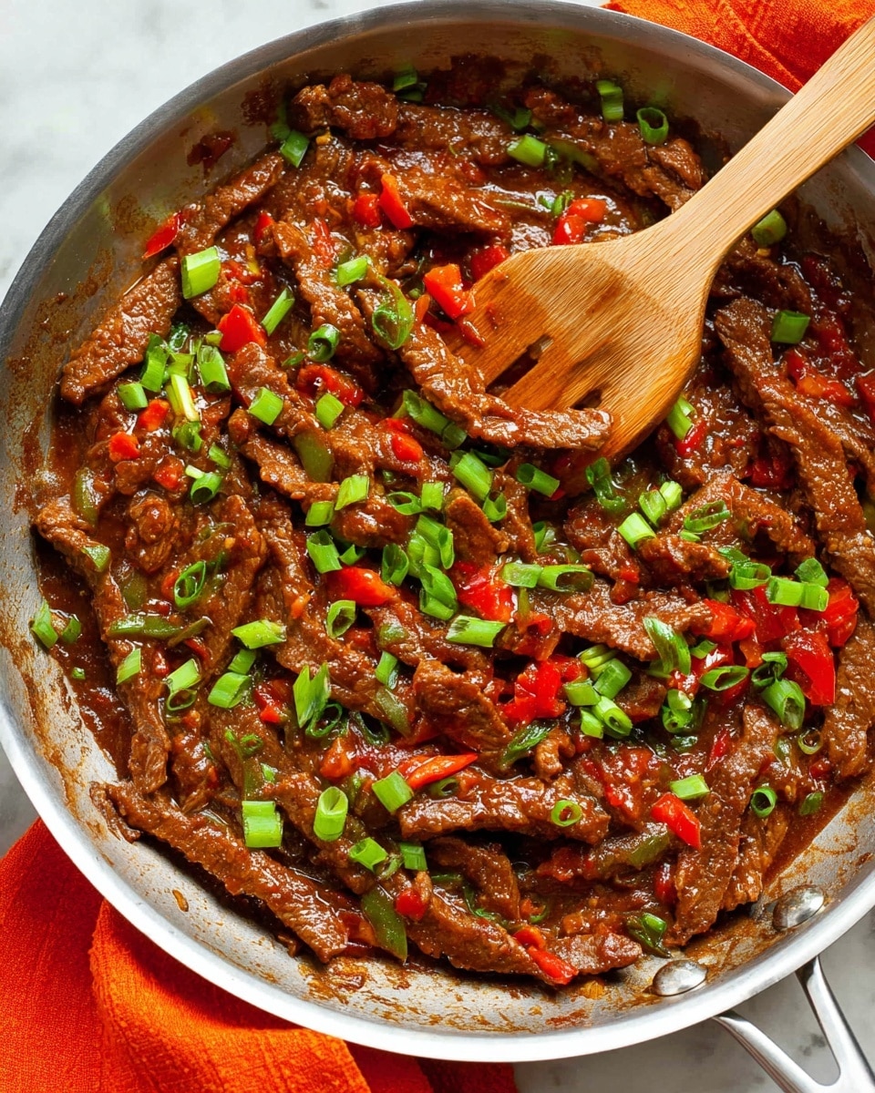 The image shows a shiny silver pan filled with cooked beef strips coated in a thick, dark brown sauce. Bright green chopped scallions are scattered on top, adding fresh color, along with small red bell pepper pieces mixing into the sauce. A wooden spatula rests inside the pan, partially stirring the beef and sauce mixture. The pan is set on a white marbled surface with a bright orange cloth partially visible underneath one side. photo taken with an iphone --ar 4:5 --v 7