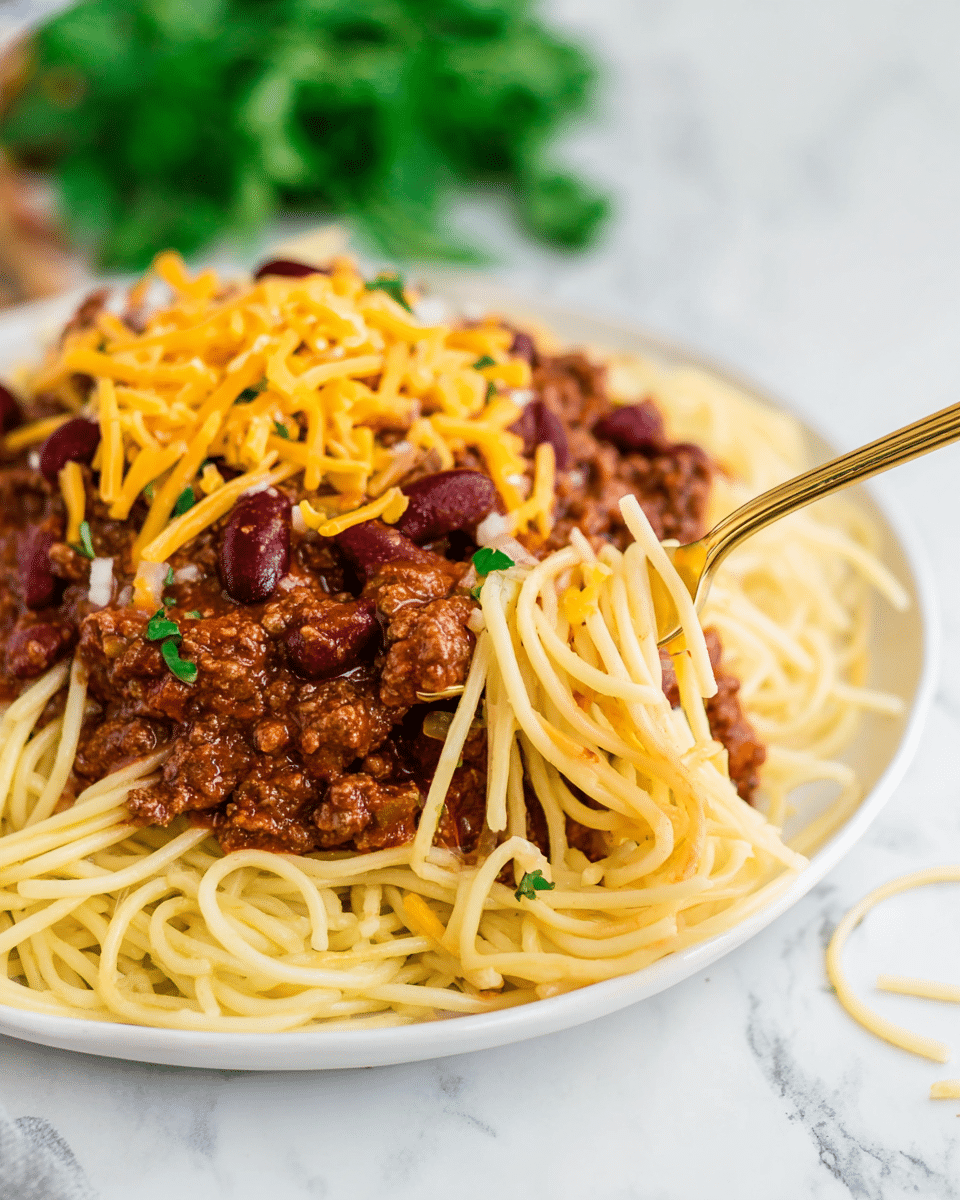 A white plate holds a serving of spaghetti with three visible layers. The bottom layer is light yellow spaghetti noodles twisted and spread across the plate's front. On top of the noodles is a thick layer of dark brown cooked ground meat sauce with a slightly chunky texture. Scattered within the meat sauce are dark red kidney beans and small pieces of diced white onions. Bright orange shredded cheese is sprinkled over the top layer of beans and onions. A gold fork is stuck into the meat sauce near the plate's center, lifting some sauce and noodles. The plate sits on a white marbled surface with blurred green herbs in the background. Photo taken with an iphone --ar 4:5 --v 7